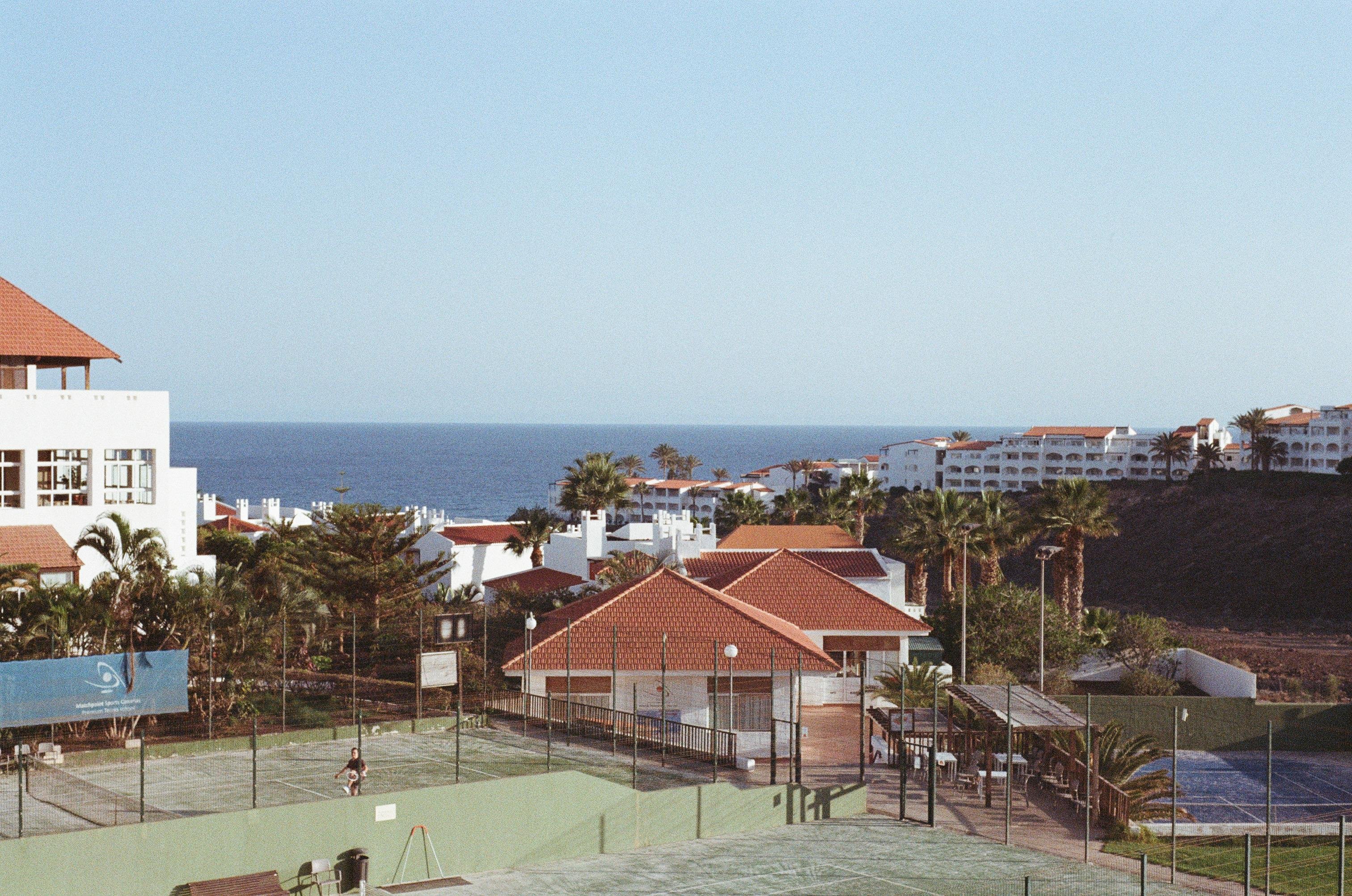 The image shows a coastal area with several buildings featuring red-tiled roofs. There's a tennis court in the foreground, and the ocean is visible in the background. The area is lined with palm trees, suggesting a resort or residential complex by the sea.