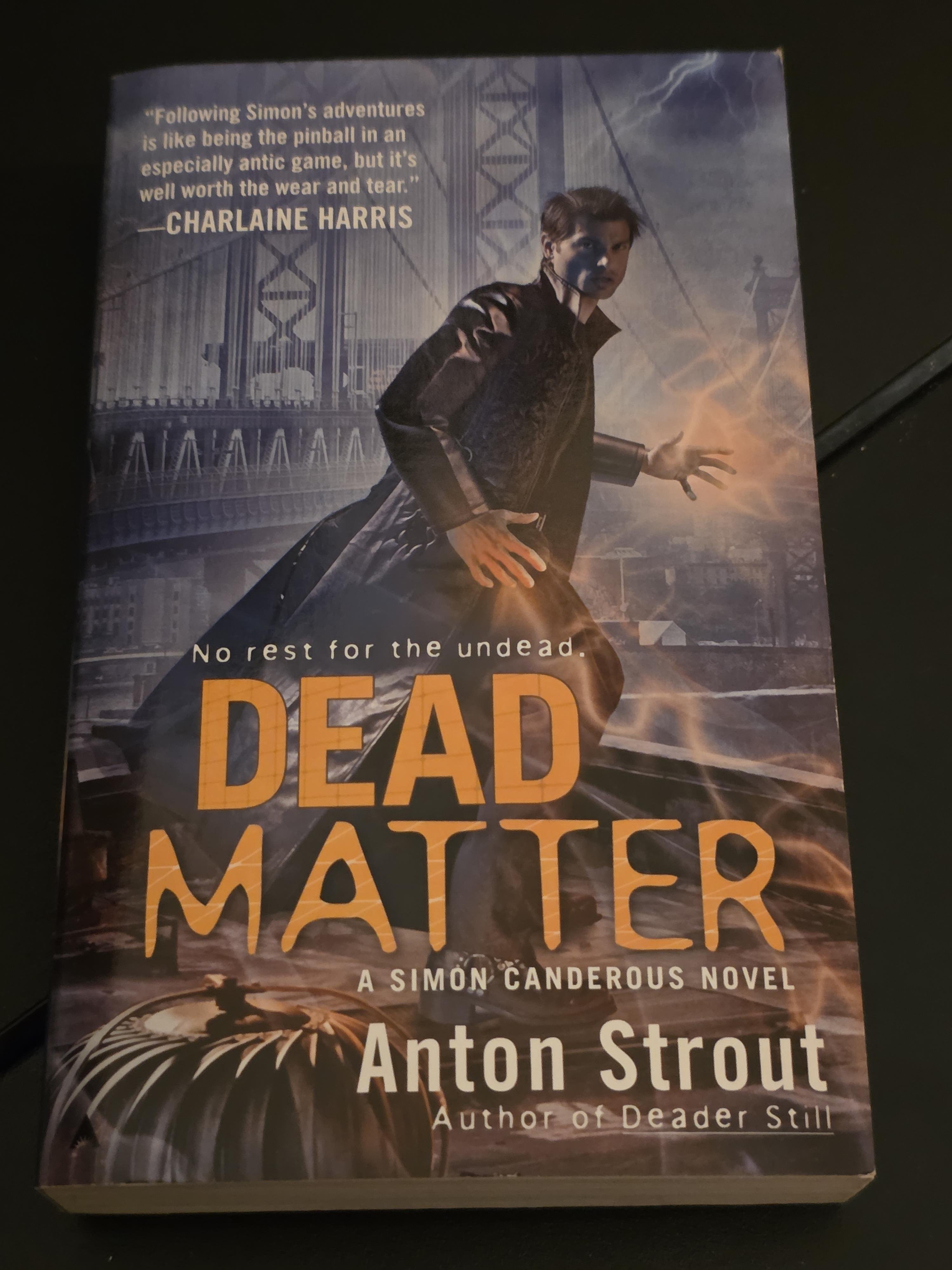 The cover of the urban fantasy novel "Dead Matter" by Anton Strout features a man in a long, dark coat standing on a suspension bridge, such as the Manhattan Bridge, under a stormy sky. His hand glows with an orange, magical energy, and bolts of lightning strike in the background. A quote from author Charlaine Harris at the top praises the protagonist Simon Canderous's "antic" adventures. This is the third book in the Simon Canderous series, where the titular hero works for New York's Department of Extraordinary Affairs, using his psychometric powers to solve supernatural crimes involving ghosts and the undead. The bold, yellow title "DEAD MATTER" sits above the tagline "No rest for the undead."