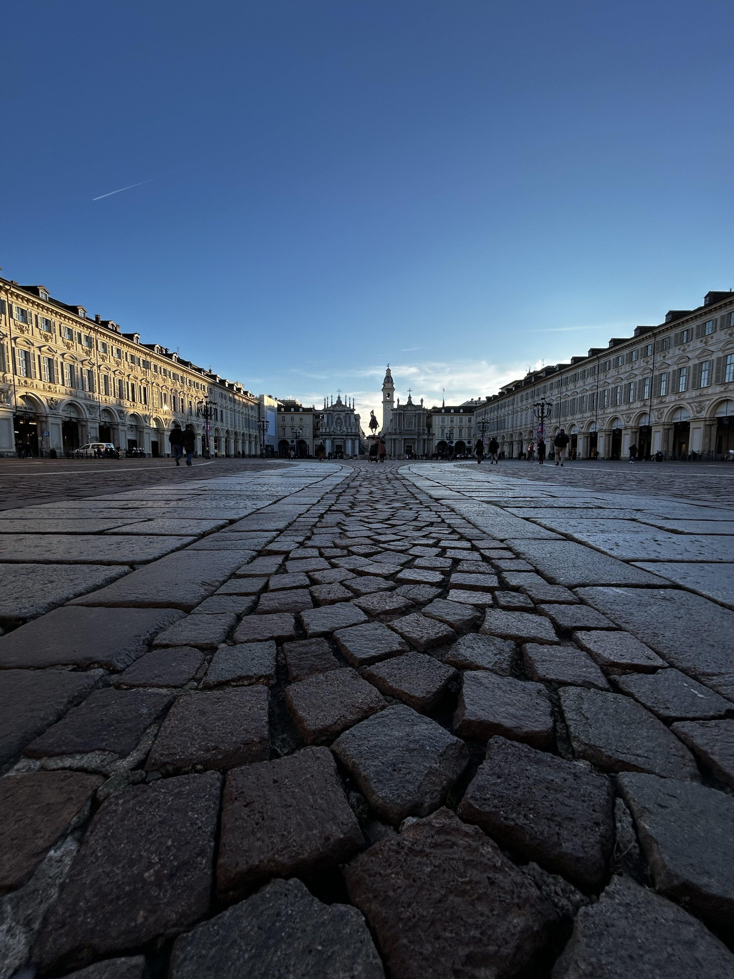 The image depicts a wide, cobblestone plaza with symmetrical buildings on both sides, leading to an ornate church or cathedral in the background. The angle is from a low perspective, emphasizing the cobblestones in the foreground. The sky is clear and blue, indicating daylight. This appears to be a historic or prominent European square.