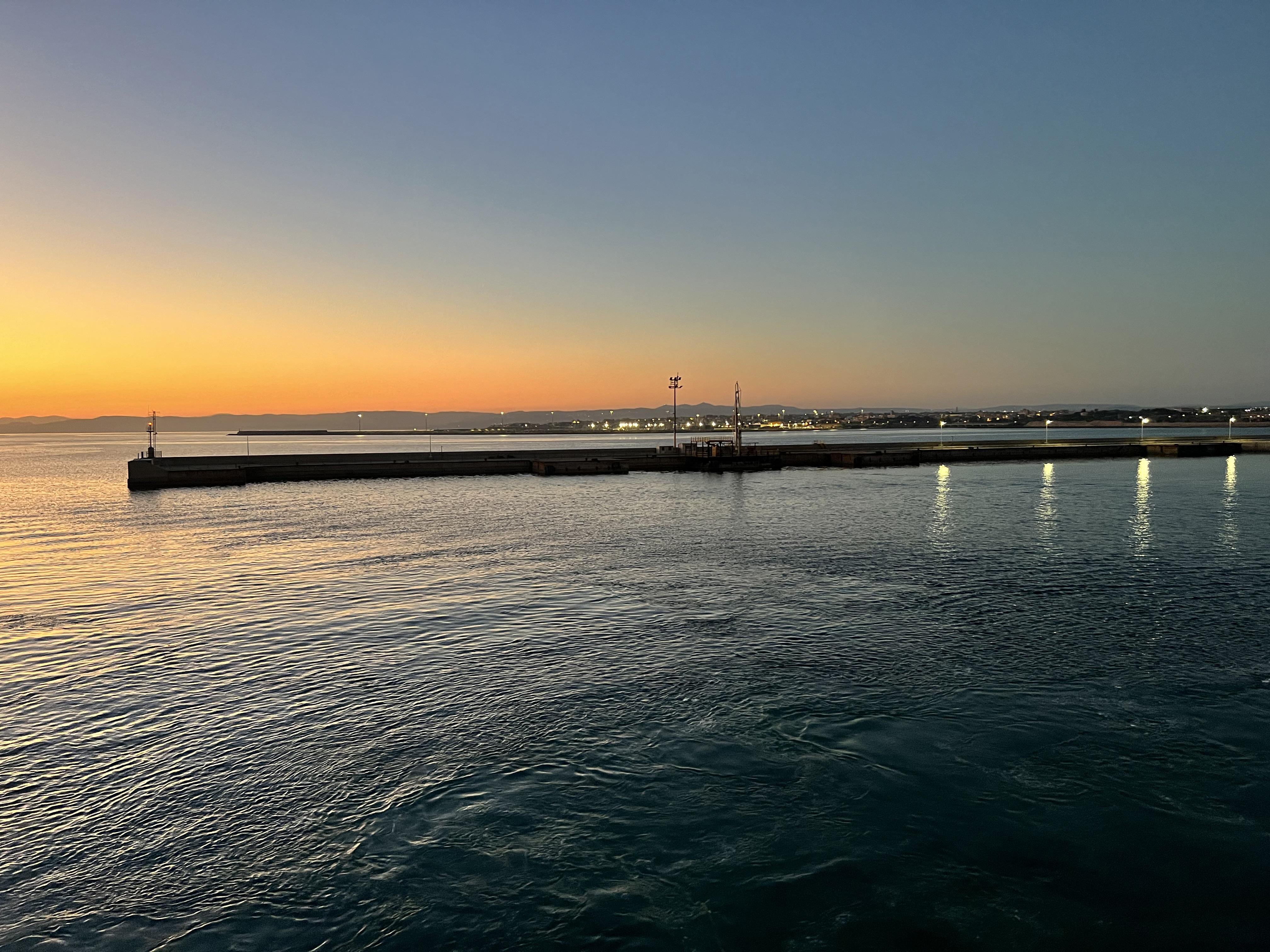 Vista pittoresca di un porto all'alba. Il cielo presenta una sfumatura dal blu all'arancione, indicando che il sole sta sorgendo. Ci sono luci lungo una diga o un molo, e l'acqua calma riflette queste luci. All'orizzonte si vedono terre o edifici lontani, contribuendo a un'atmosfera tranquilla e scenografica.