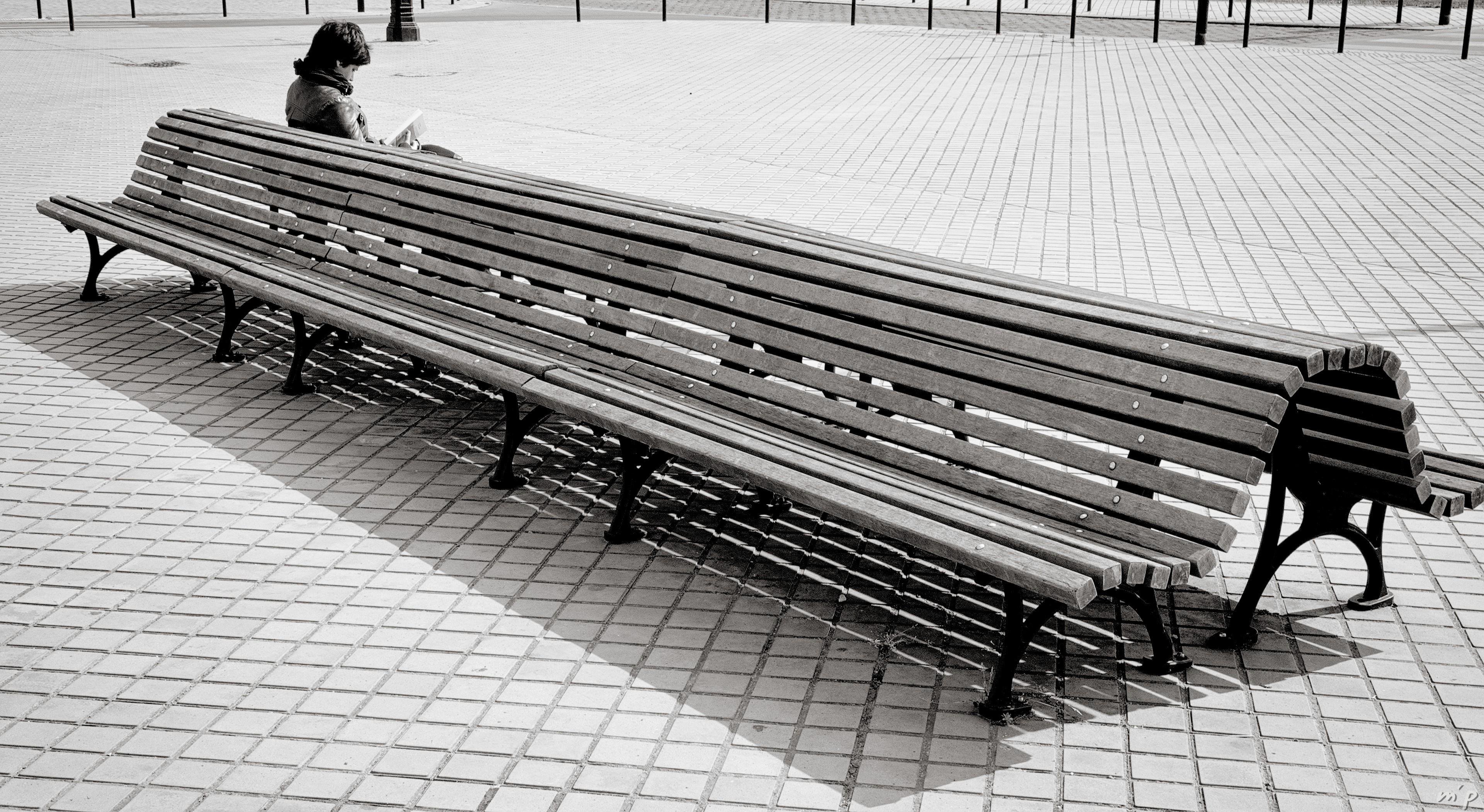 🇬🇧 Horizontal black and white photo showing a sunny square with a double back-to-back bench of wooden slats taking up the entire diagonal of the image with at its end, at the top of the image, a young woman reading a book.

///

🇫🇷  Photo noir et blanc horizontale montrant une place ensoleillée avec un banc double dos à dos de lattes de bois prenant toute la diagonale de l'image avec à son bout, en haut de l'image, une jeune femme brune lisant un livre.