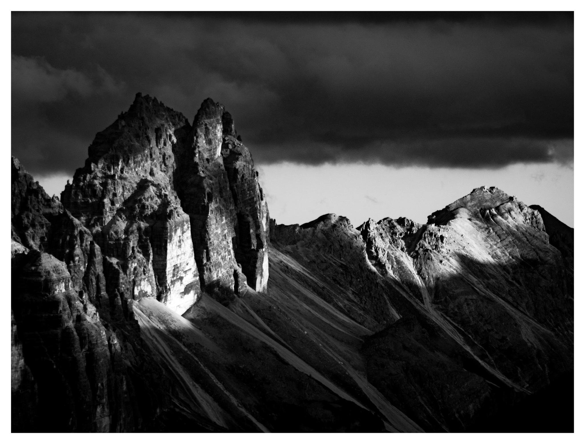 Black-and-white landscape of jagged mountain peaks, with a tall central rock spire lit by sunlight against deep shadows; dark storm clouds stretch across the sky above a ridgeline sloping to the right.