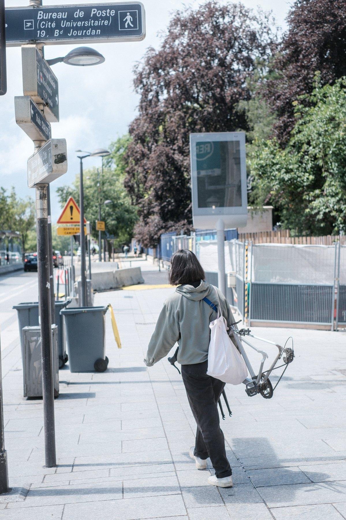 Photo verticale. Une jeune femme brune au cheveux en carré, de dos, hoodie vert pâle, jean noir, basket blanches, sac tote blanc sur l’épaule droite, marche en tenant un cadre de vélo sans roue de la main droite