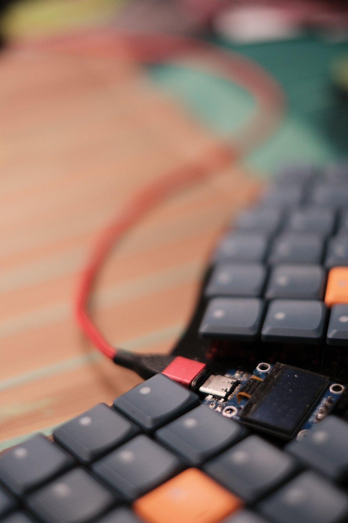 Photo verticale. Gros plan sur un clavier ergonomique aux touches bleu/gris sans lettres, juste un point blanc comme marquage. Parmis elles, 2 touches oranges. Il y a une partie sans touche au centre, occupée par un écran et une prise USB rouge et noir, suivi par un câble rouge. La profondeur de champs est très fine, ne laissant que la prise et l’écran nets, le reste dans le bokeh