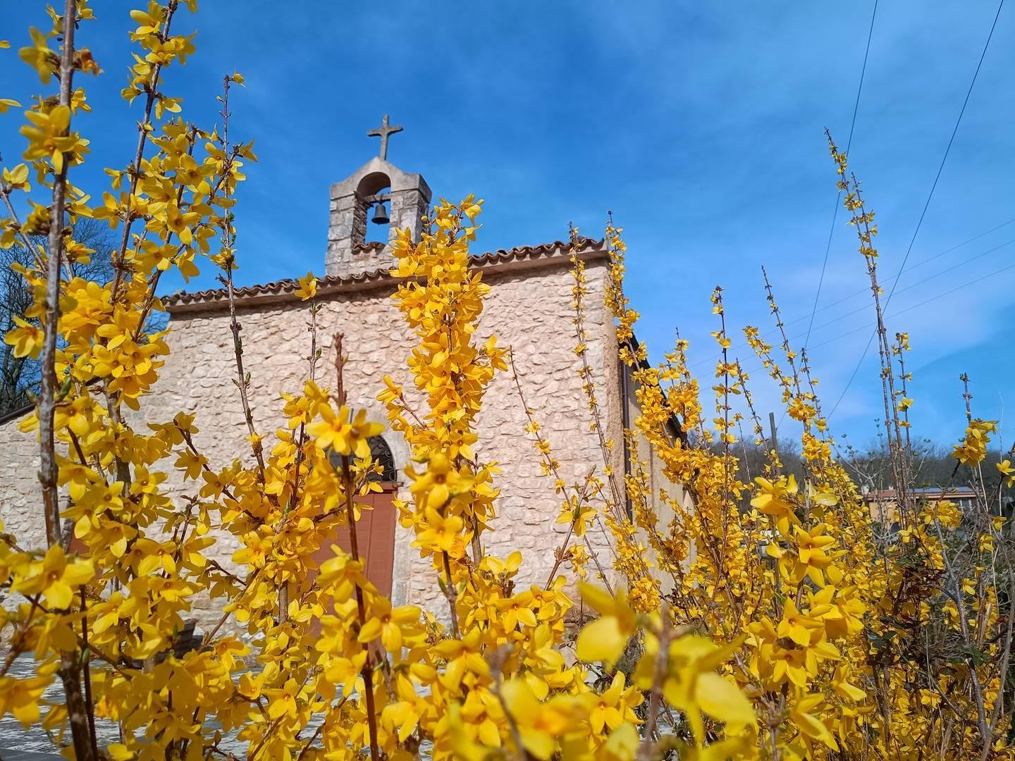 Chiesa di campagnia, con un piccolo campaniele sormontato da una croce, tra il giallo dei fiori in primo piano e il blu del cielo.