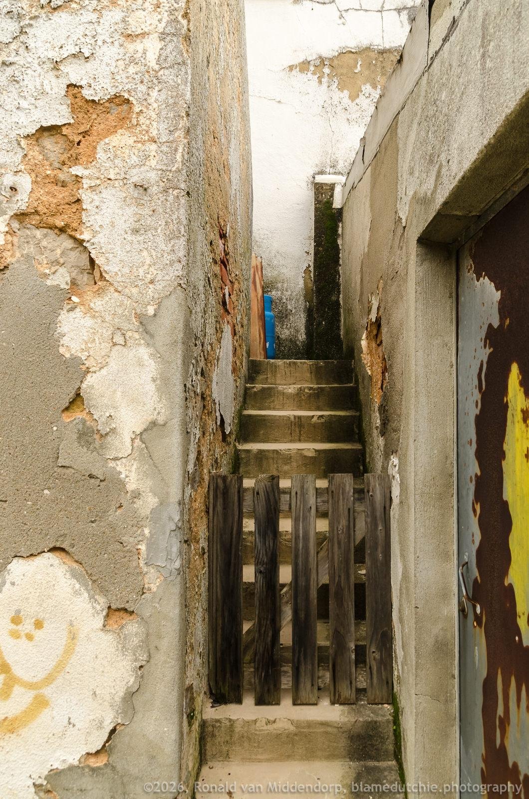 Narrow outdoor passage between two weathered walls with peeling plaster, leading up a short flight of concrete steps blocked by a small wooden fence; rusted metal door on the right and a blue gas bottle partially visible at the top.