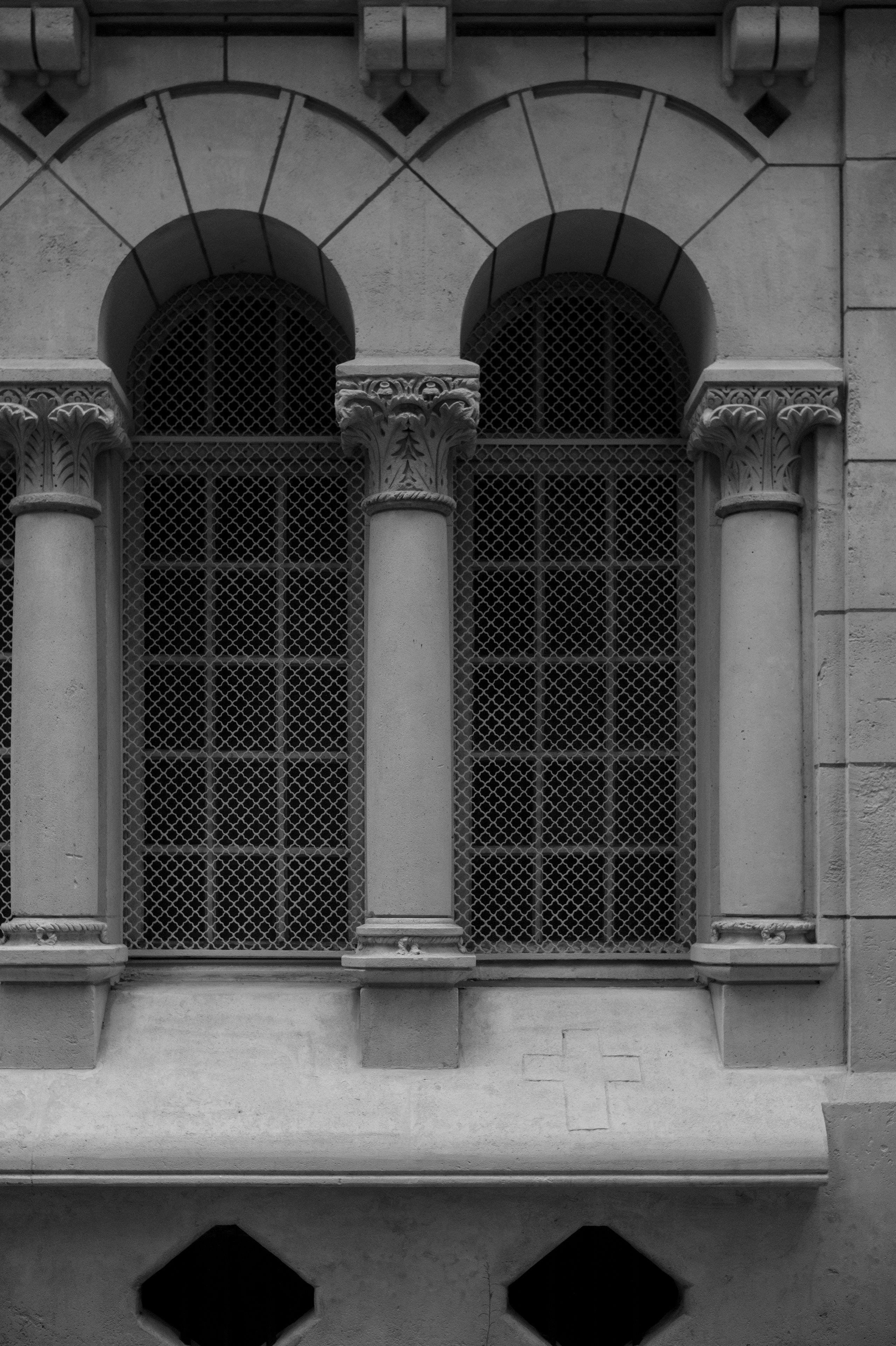 Photo verticale en noir et blanc. 2 Fenêtres encadrées par une décoration avec des colonnes et des arches. Sur un espèce d’appui de fenêtre en pente vers la caméra, on distingue une croix gravée comme un graffiti