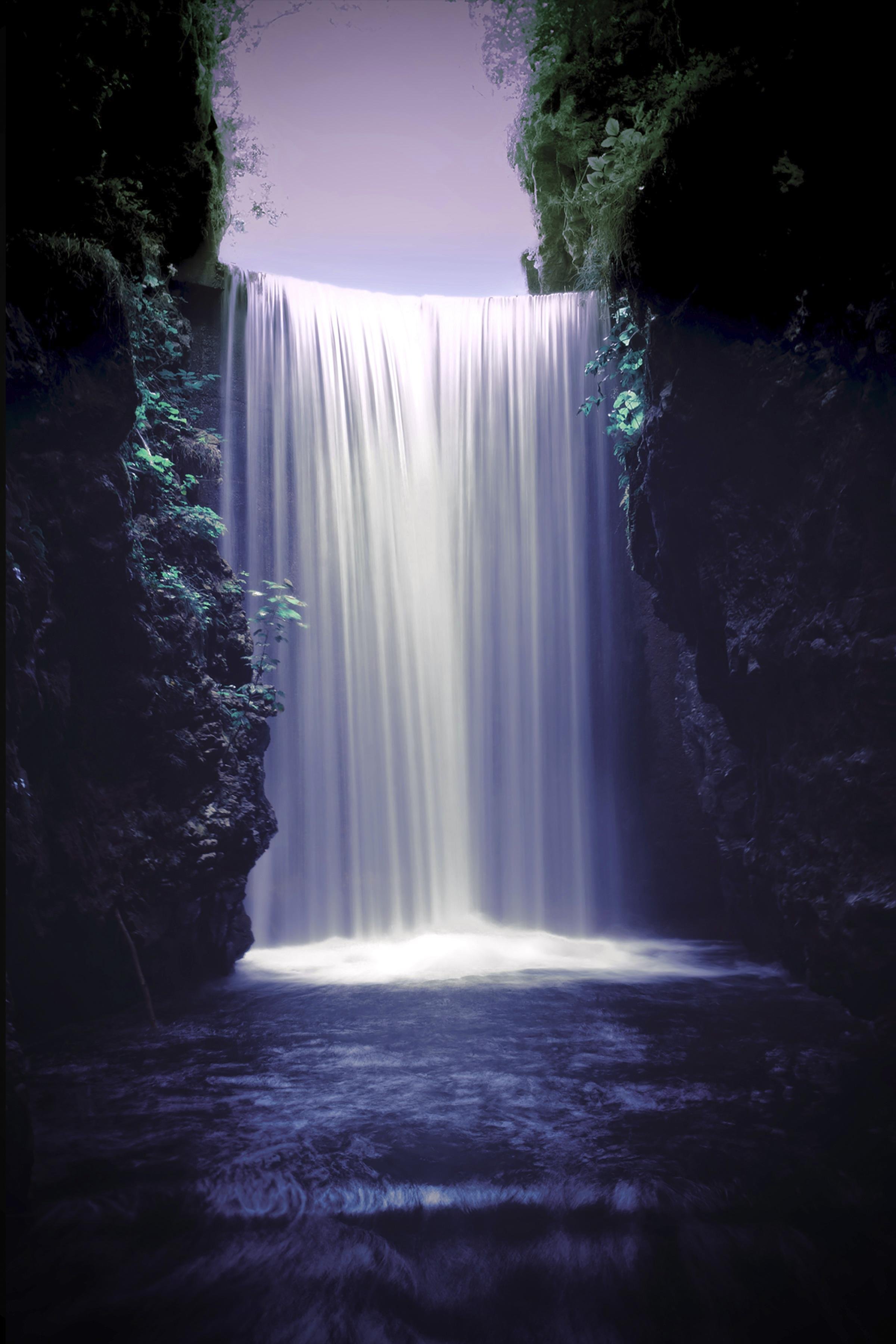 Tall waterfall dropping in a smooth white curtain between dark, mossy rock walls into a shadowed pool, with small green plants clinging to the cliff edges and pale sky visible above the falls.