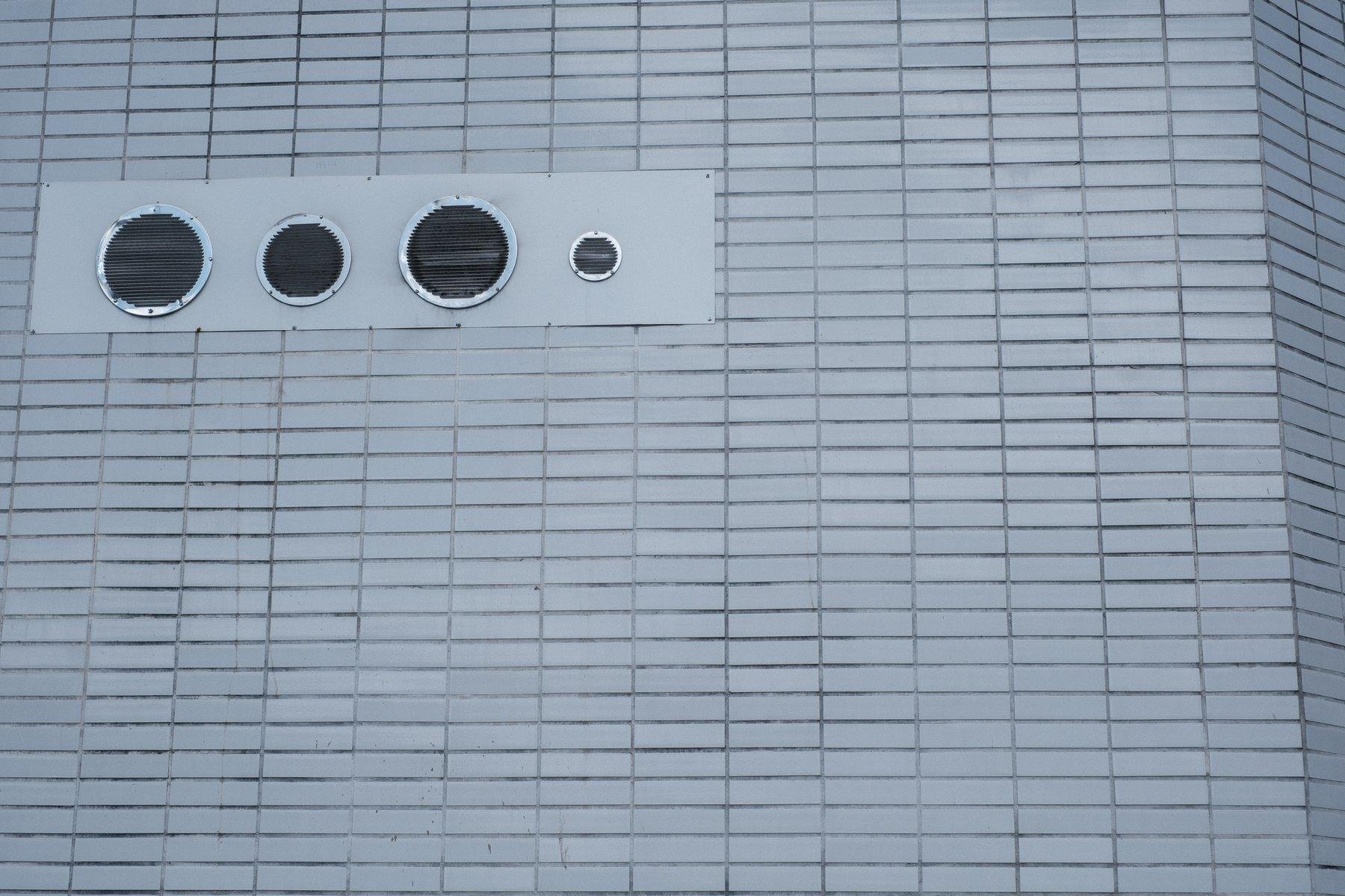 Photo horizontale. Sur un mur fait de petite tuile rectangulaire horizontales grises, une partie est sans tuile, gris un peu plus clair, avec 4 évacuations avec des lames protectrice, une grosse, une moyenne, une autre grosse, et une petite