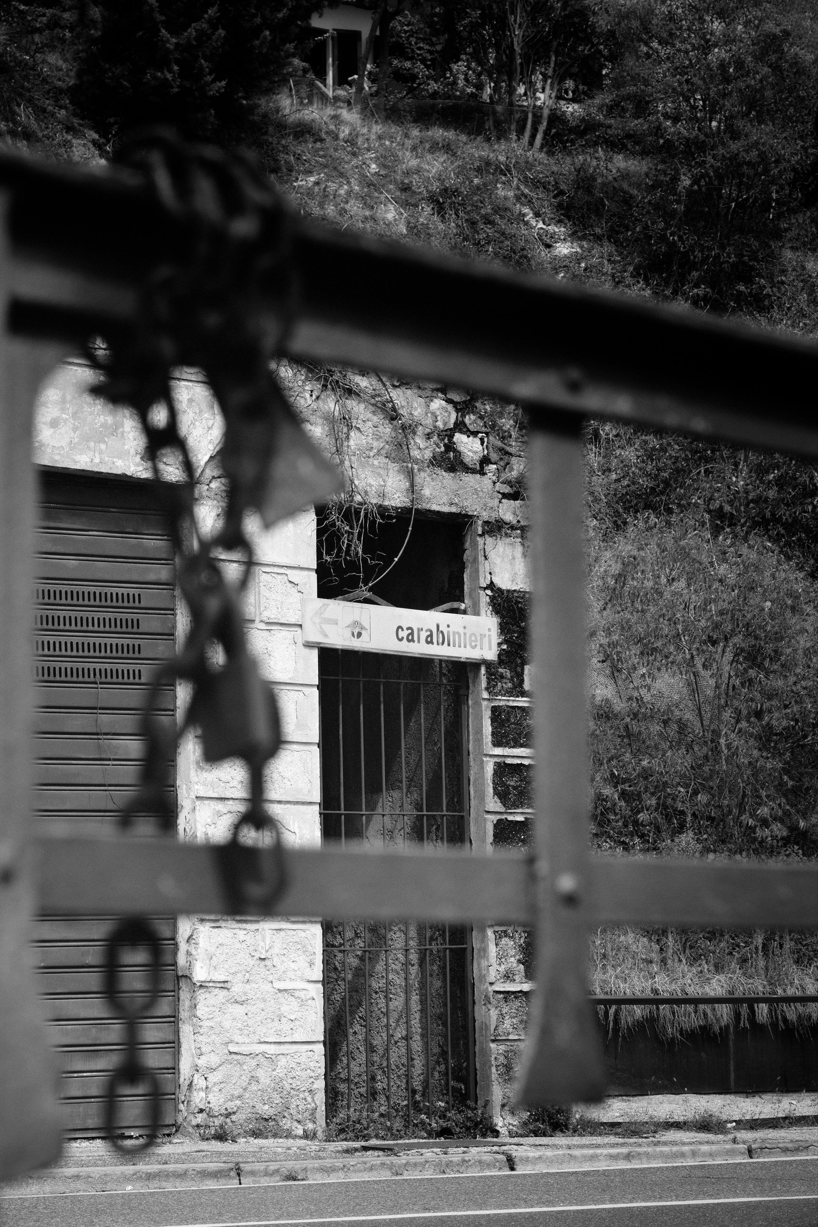 Blurred metal bars and a rusty chain with a padlock in the foreground frame an old stone building with a closed brown shutter and a barred doorway; a heavily weathered sign above the doorway reads “carabinieri” with a left-pointing arrow.