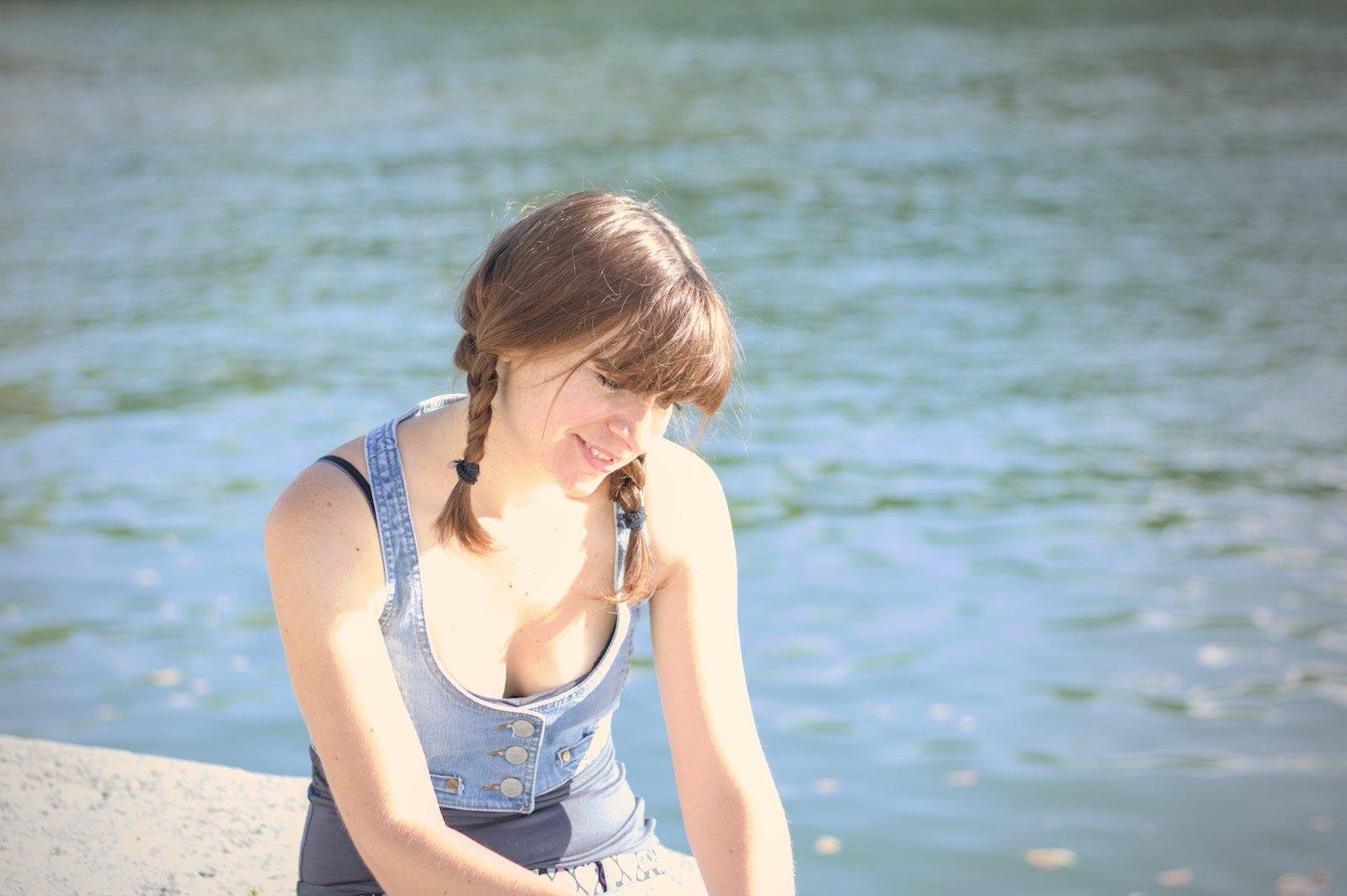 Une jeune femme brune coiffée avec des nattes, assise de 3/4, regardant en bas, devant elle, les mains jointes (hors cadre) un sourire aux lèvres, éclairée par le soleil, la Seine en fond. Elle porte un débardeur bleu marine, et un gilet en denim par dessus