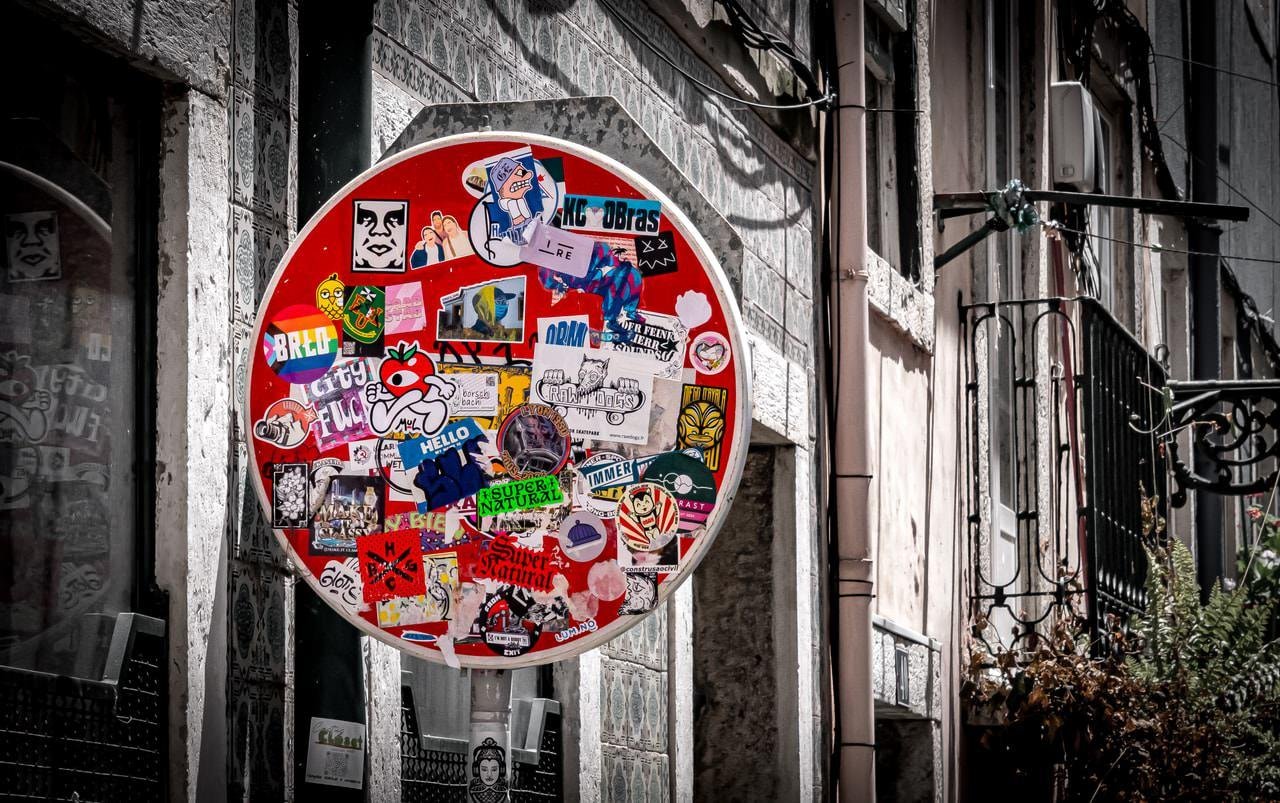 Red circular sign covered with various colorful stickers, featuring faces, logos, and slogans. Background includes a weathered stone wall and a building facade with windows and metal railings, situated in an urban setting.