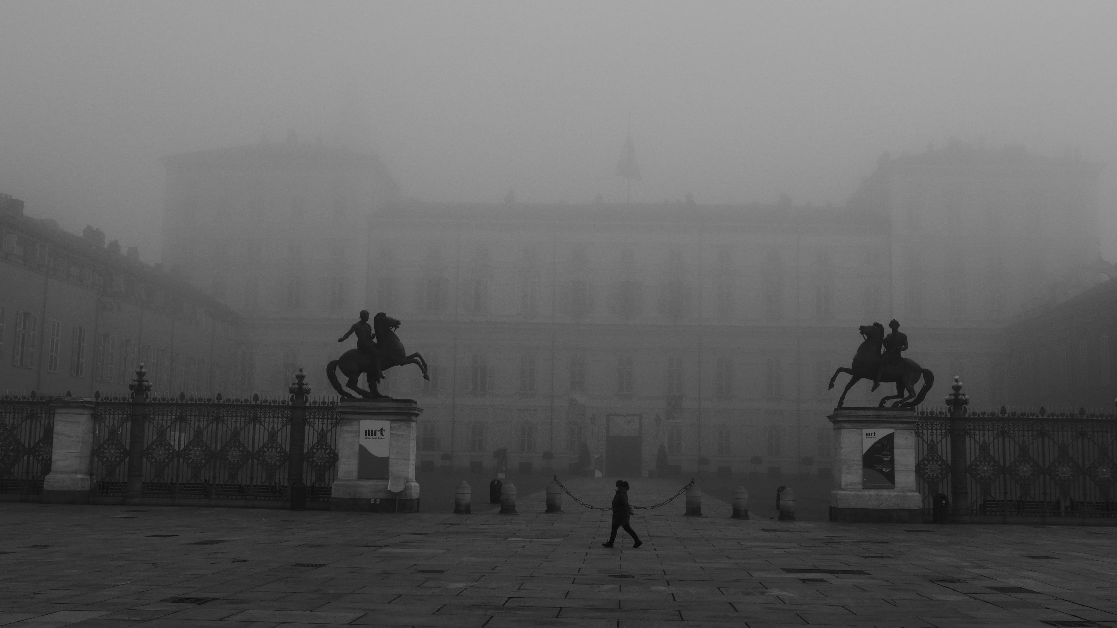 The image depicts a foggy scene of a grand building with large gates in front and statues of mounted figures on either side. A person is walking across the plaza in the foreground.