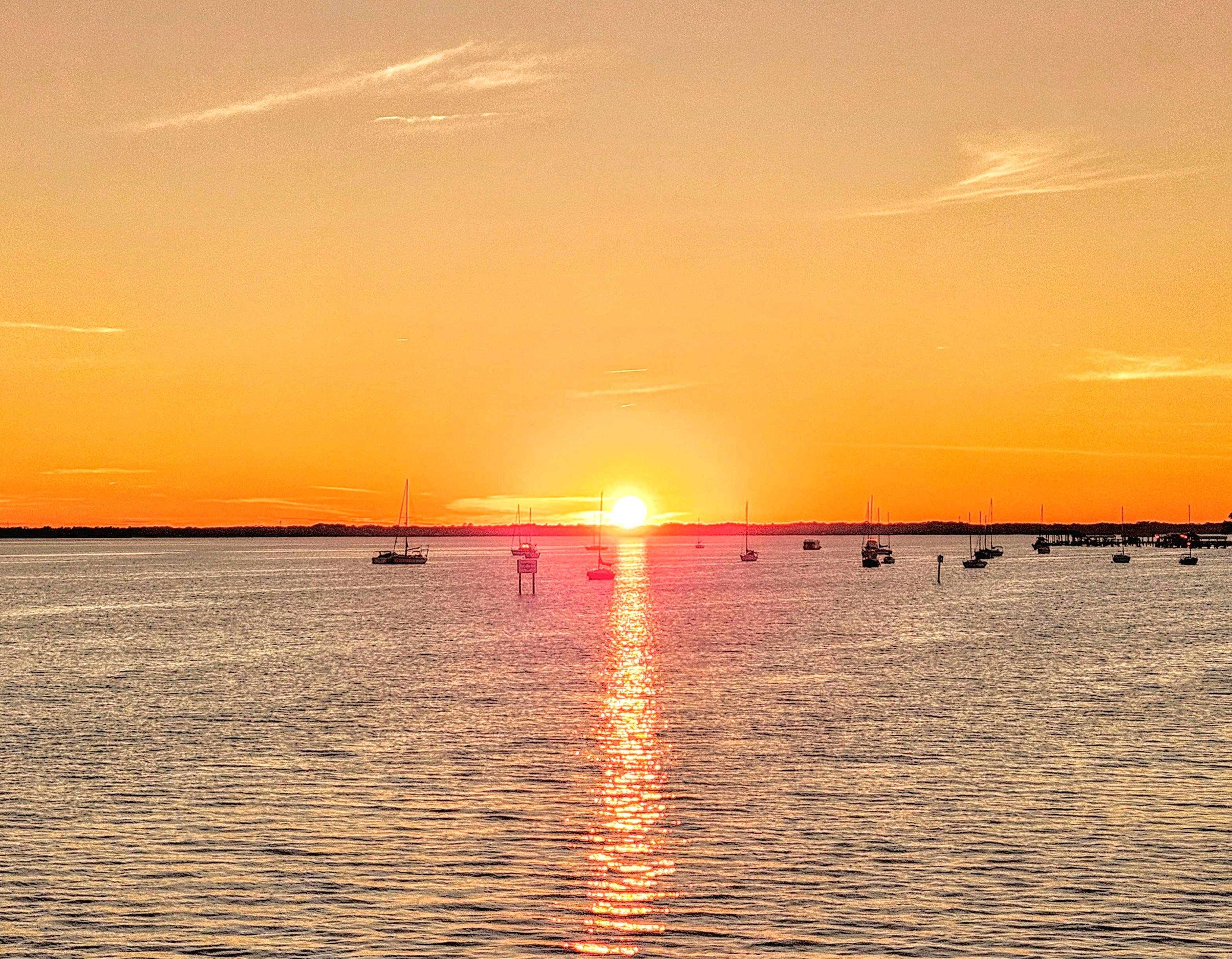 Sunset over a calm sea with several small boats scattered across the water. The sky is a gradient of warm orange and yellow hues, with the sun near the horizon casting a bright reflection on the water's surface. Thin clouds are visible in the sky, adding texture to the scene.