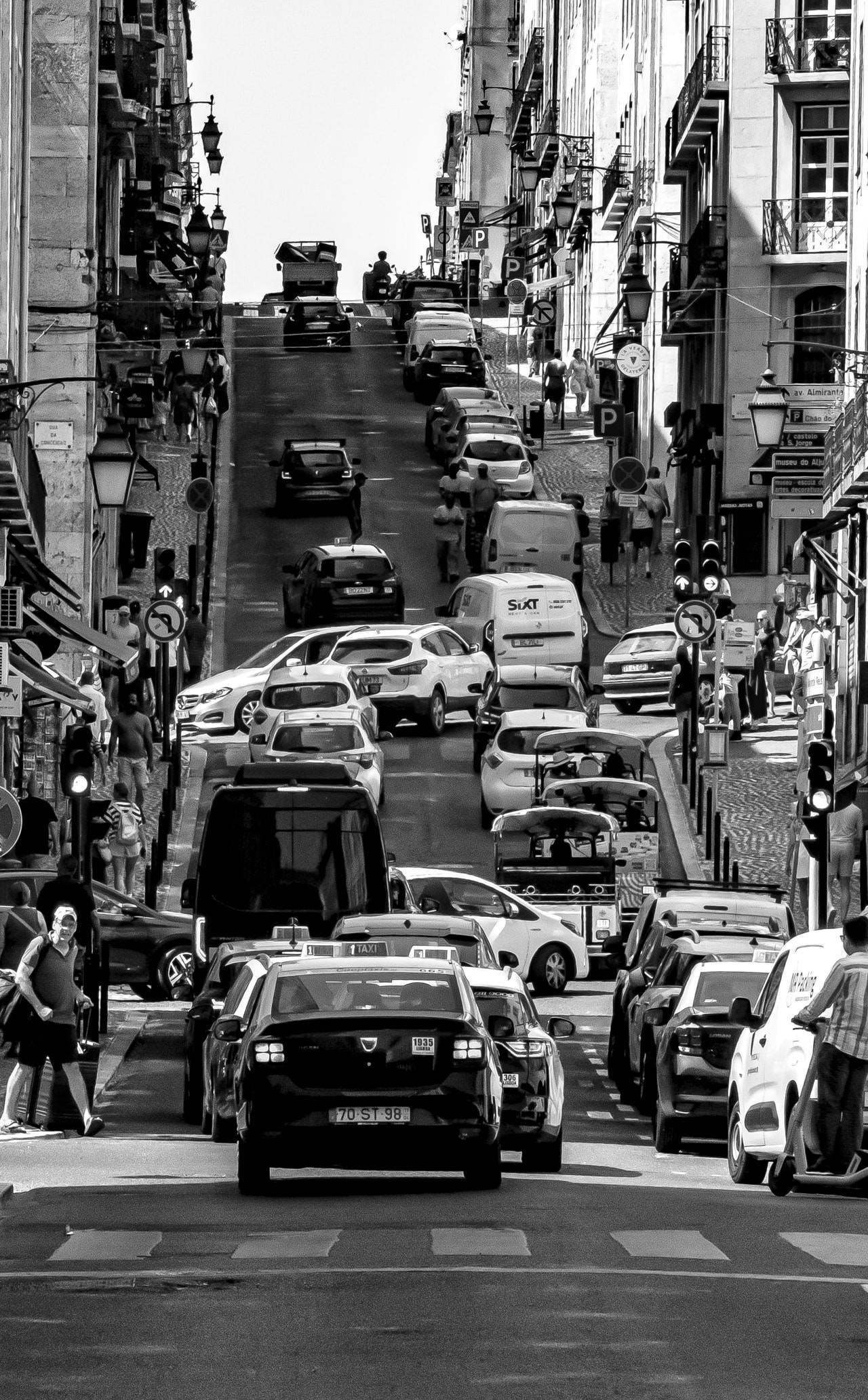 Narrow, busy street with steep uphill slope crowded with cars, vans, and taxis. People walk along sidewalks, with parked scooters and motorcycles visible. Buildings with balconies and street lamps line both sides. Traffic signs and signals scattered throughout, adding to the urban setting. Black and white tones emphasize shadows and contrasts.