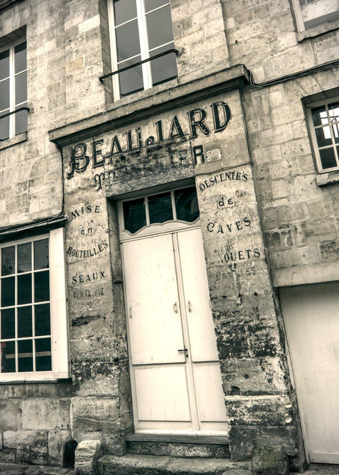 Photo format portrait en sépia, montrant la façade d’une vieille cave à vin, dont l’enseigne délavée par le temps dit « Beaujard », en dessous, c’est trop abîmé pour être lu, et sur chaque côté de la porte blanche, « Mise en bouteilles » « Descentes de caves » « Seaux » et d’autres lettres effacées.