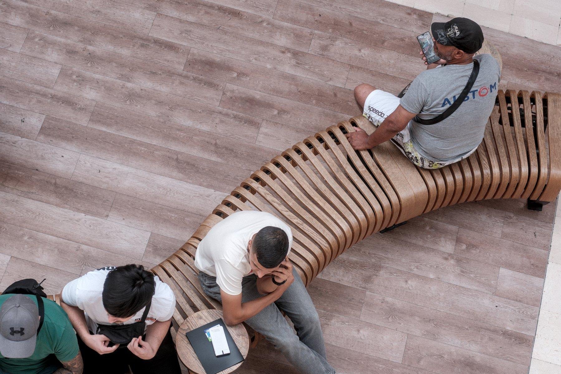 Photo horizontale. Vue de dessus, un banc fait de lattes de bois dans le sens des jambes, formant un serpentin dans le cadre, avec des gens assis dessus. Dans une galerie marchande