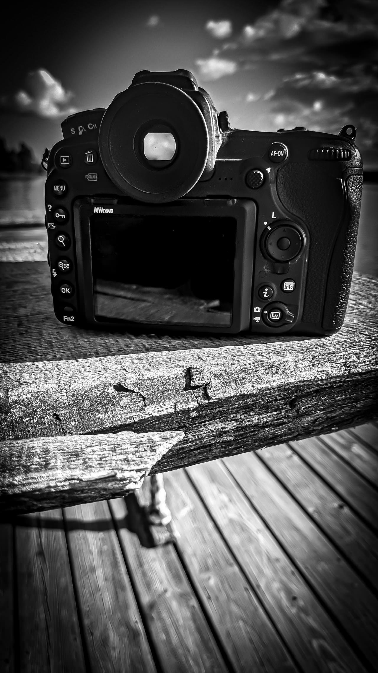Black and white photo of a Nikon camera resting on a weathered wooden surface. The camera is positioned with its back facing the viewer, displaying various buttons and a viewfinder. The background includes a blurred scenery with clouds in the sky, indicating an outdoor setting.
