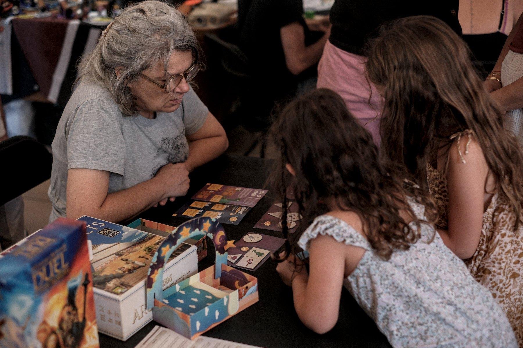 Photo horizontale. À une table avec une nappe noire, une partie de jeu de société avec une dame au cheveux blanc, t-shirt gris clair, accoudée à la table devant un livret de règles et en face, 2 petites filles de 3/4 dos, brune et châtain, en robe d’été, bleu/blanc et marron/blanc
