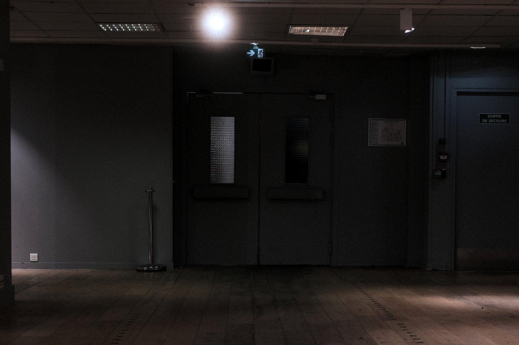Photo horizontale. Dans un endroit sombre, éclairé de ça et là, le sol en parquet, et en face, un mur gris/bleu, avec une porte de sortie de secours