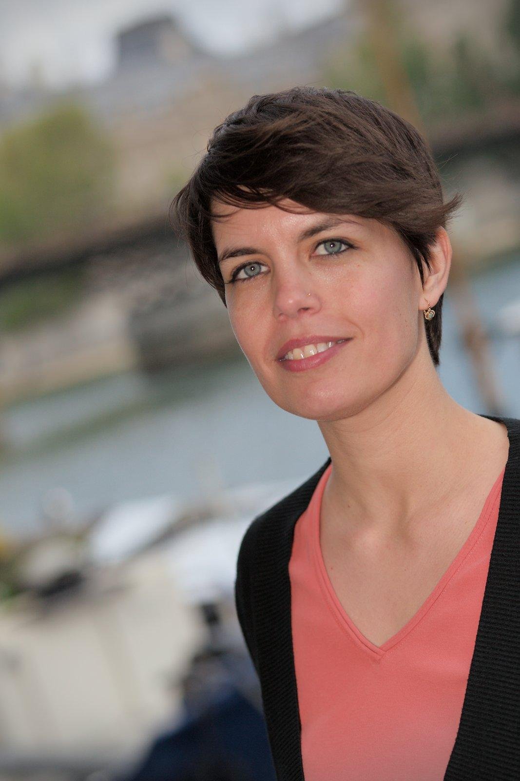 Portrait d’une brune au cheveux courts souriante, habillée avec un haut rouge/orange et un gilet noir