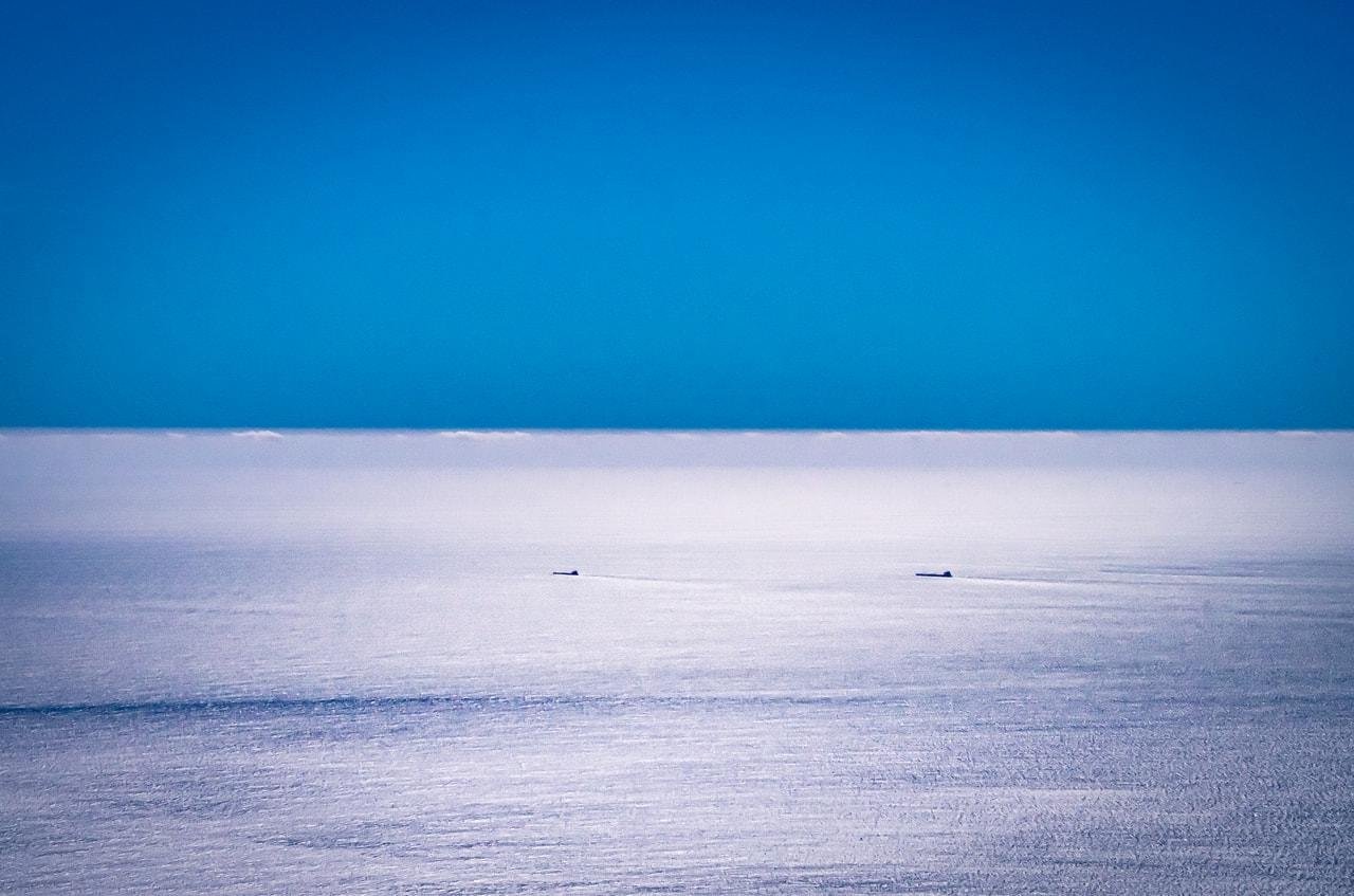 The image shows a calm ocean with a clear blue sky. There are two small ships in the distance on the horizon. The overall scene is tranquil and expansive.