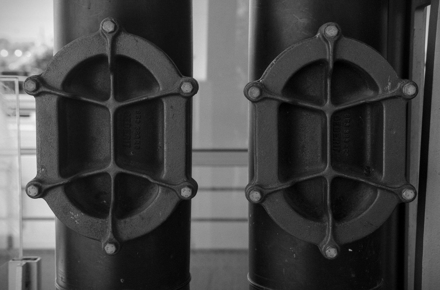 Photo horizontale en noir et blanc. Un bout de 2 tuyaux parallèles, avec une trappe de maintenance sur chaque, en forme de circuit d’athlétisme vu de dessus et solidement boulonnés par 6 écroux disposé symétriquement.