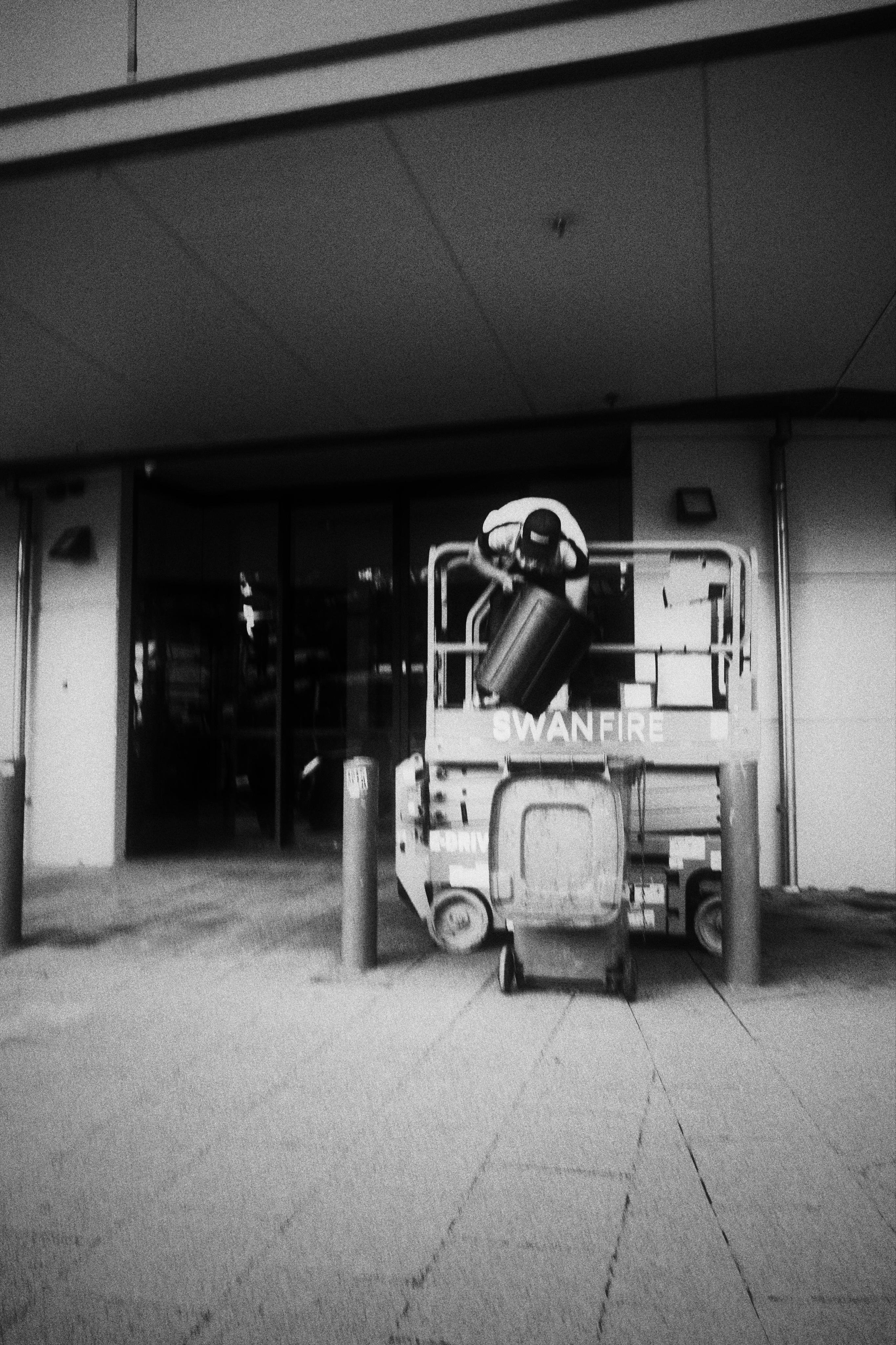 A black and white image of a scissor lift with a large rubbish bin being held by an individual over a wheelie bin. The lift is on a paved surface in front of a building with the text, “SWANFIRE” visible. The person’s face is not.
