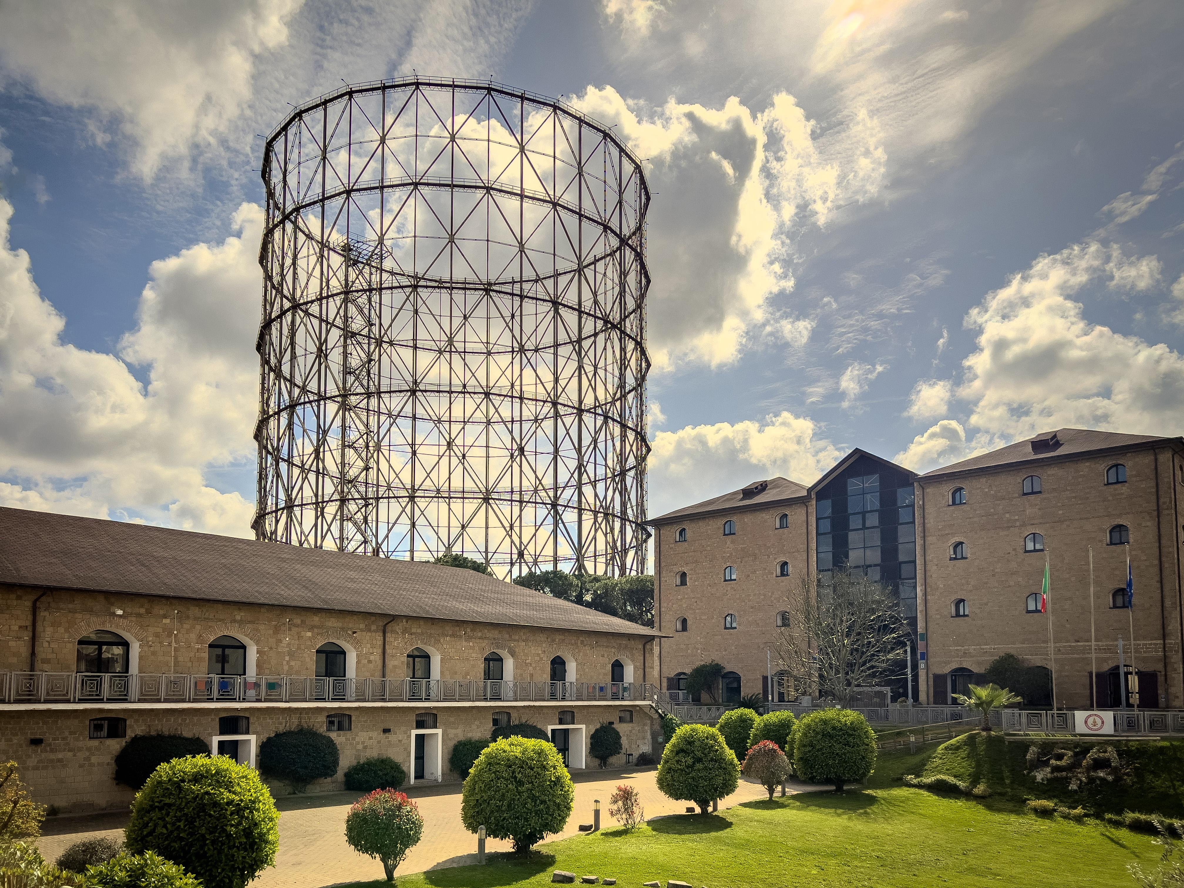 Grande struttura di un gasometro, una struttura cilindrica utilizzata per immagazzinare il gas. In primo piano, ci sono edifici in mattoni, probabilmente parte di un complesso industriale o storico, insieme a un'area verde con cespugli e alberi potati. Il cielo è parzialmente nuvoloso.