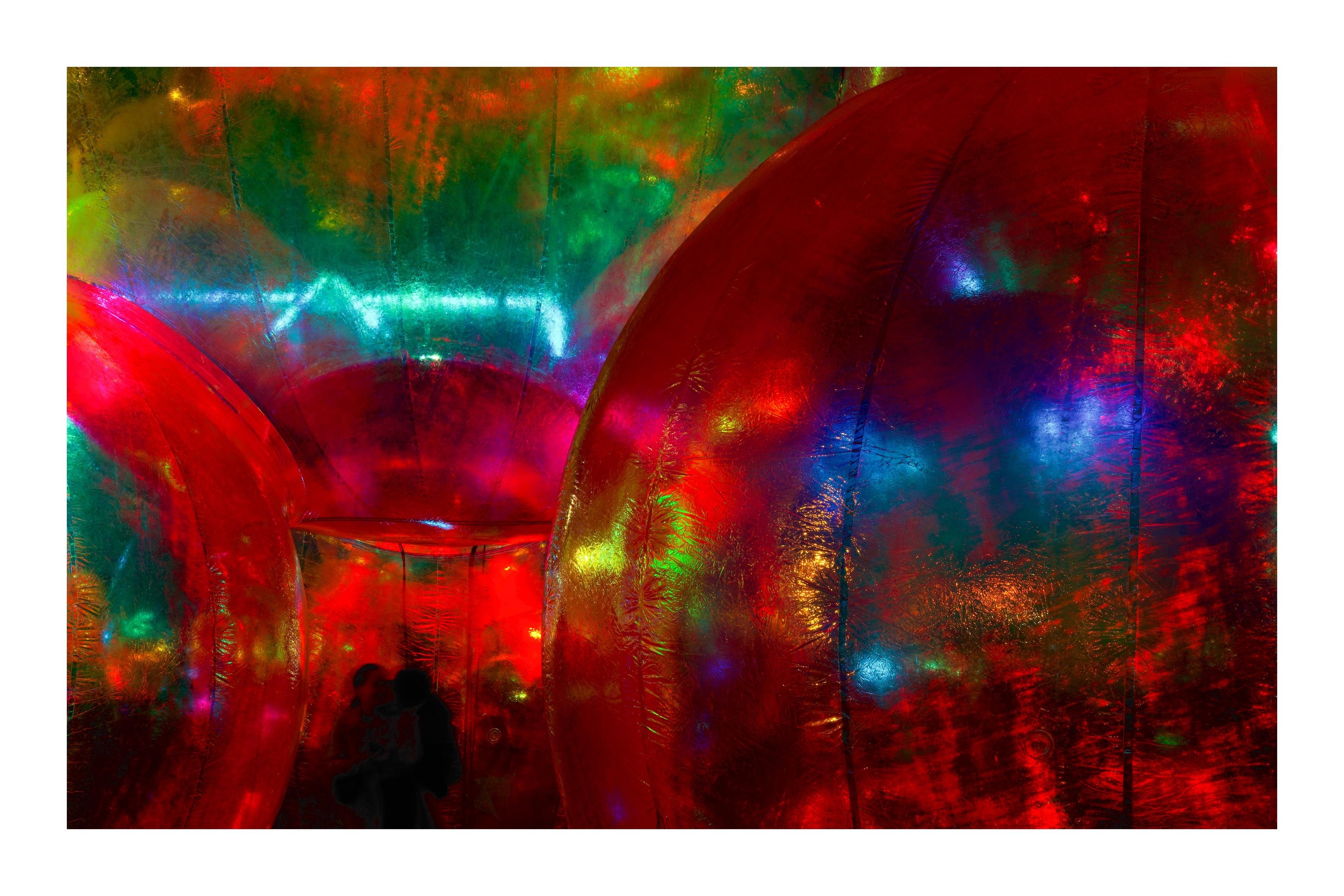 Colorful abstract scene with large, shiny, reflective spheres in shades of red, green, and blue, creating a vibrant and immersive environment. Texture on the spheres adds a shimmering effect. Two silhouetted figures are visible in the lower left corner, suggesting interaction or movement within the space.