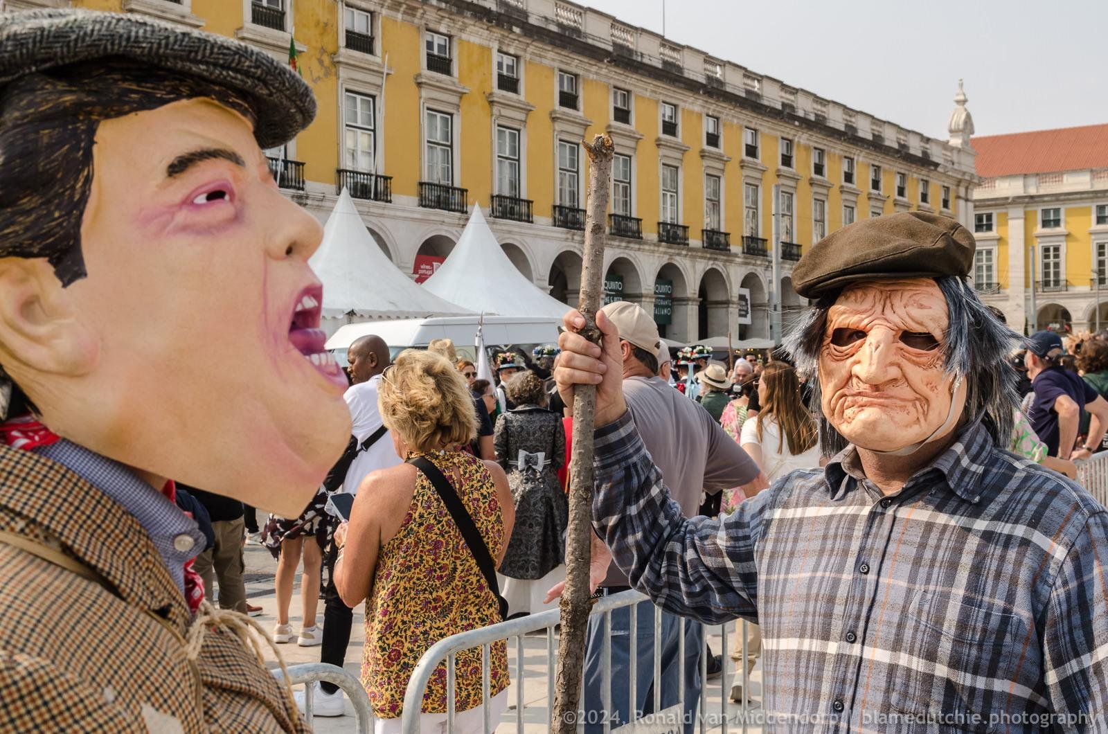 Person wearing a wrinkled old-man mask and brown cap holds a wooden staff beside a metal crowd barrier in a busy plaza; at left, a close-up of a large mask with an open mouth, with yellow arcaded buildings and white event tents in the background.