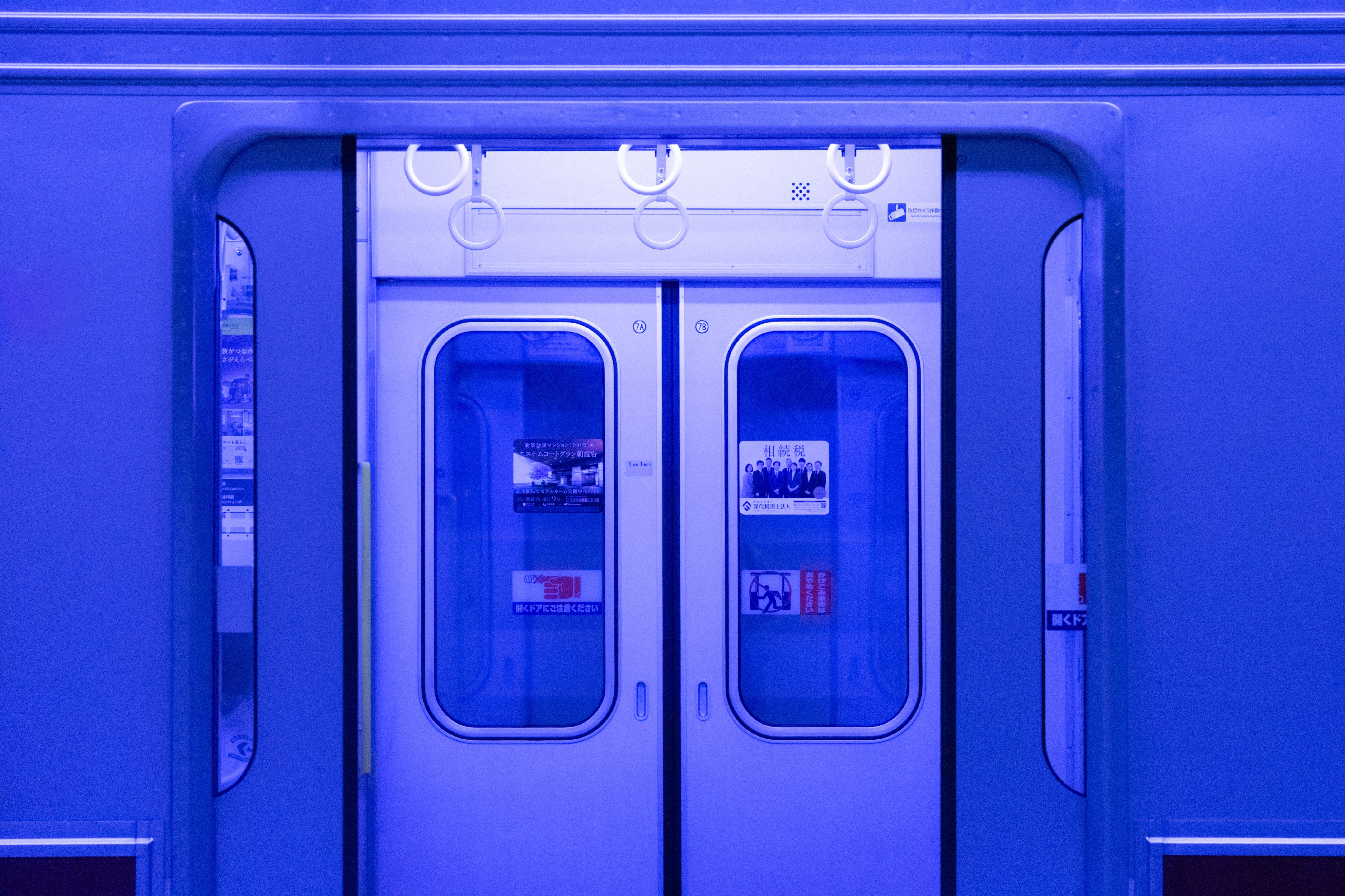 Closed sliding doors of a subway train with blue lighting. Windows on the doors display posters and signs. Overhead hand grips are visible above the doors. A metal frame surrounds the doors, and light reflects on the surface.