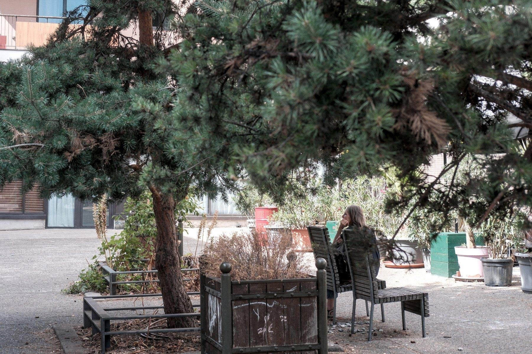 Photo horizontale. Sur une place piétonne, sous un arbre, une jeune femme est assise sur une chaise publique, perpendiculairement au dossier, de dos par rapport à la caméra, accoudée à son sac posé sur ses jambes, et tenant son menton de la main gauche
