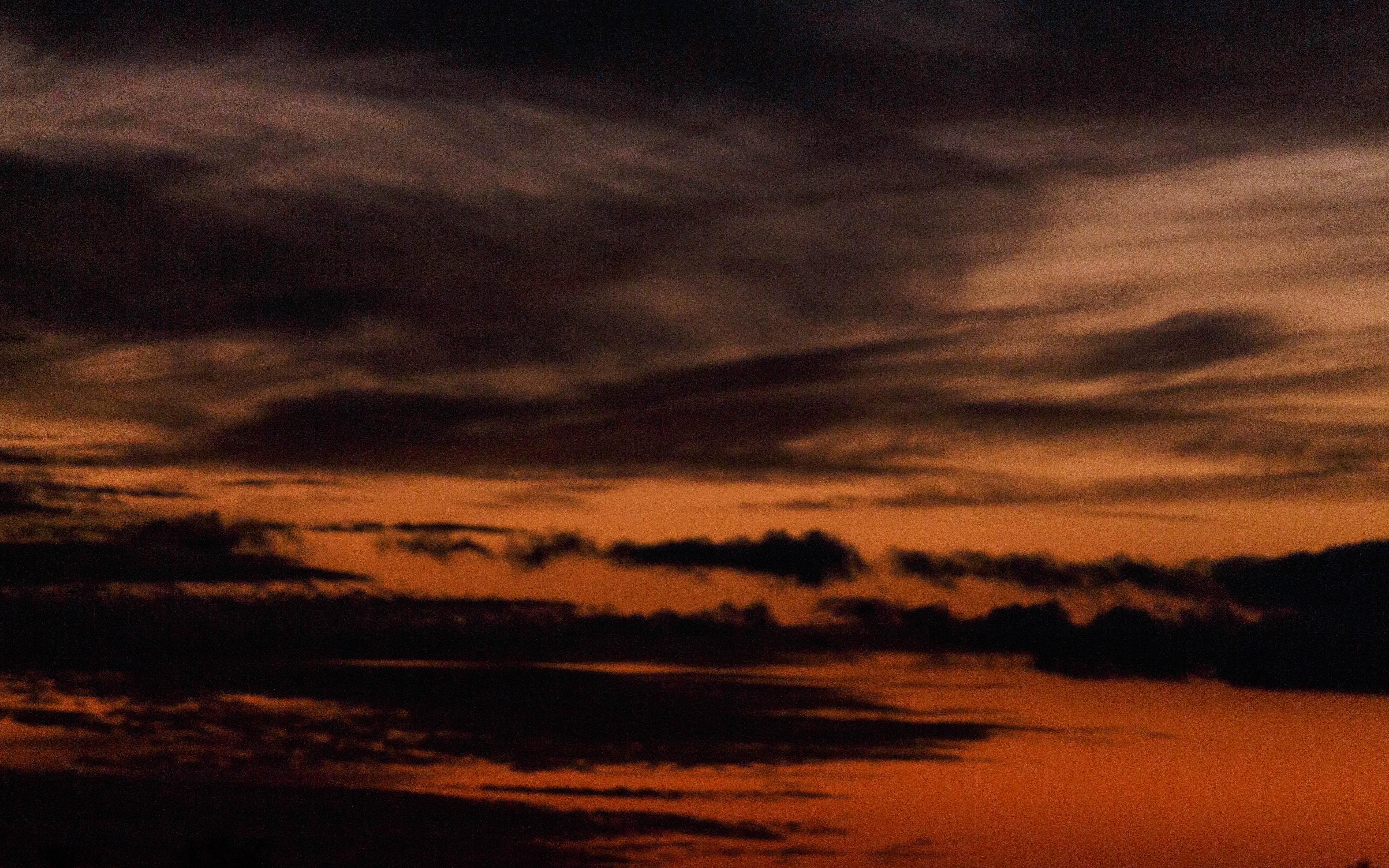 Orange and dark gray sunset sky with scattered, wispy clouds creating a layered effect. The lower part of the image shows faint silhouettes of distant clouds against the glowing horizon, highlighting the transition from glowing orange near the horizon to deeper gray tones in the upper sky.