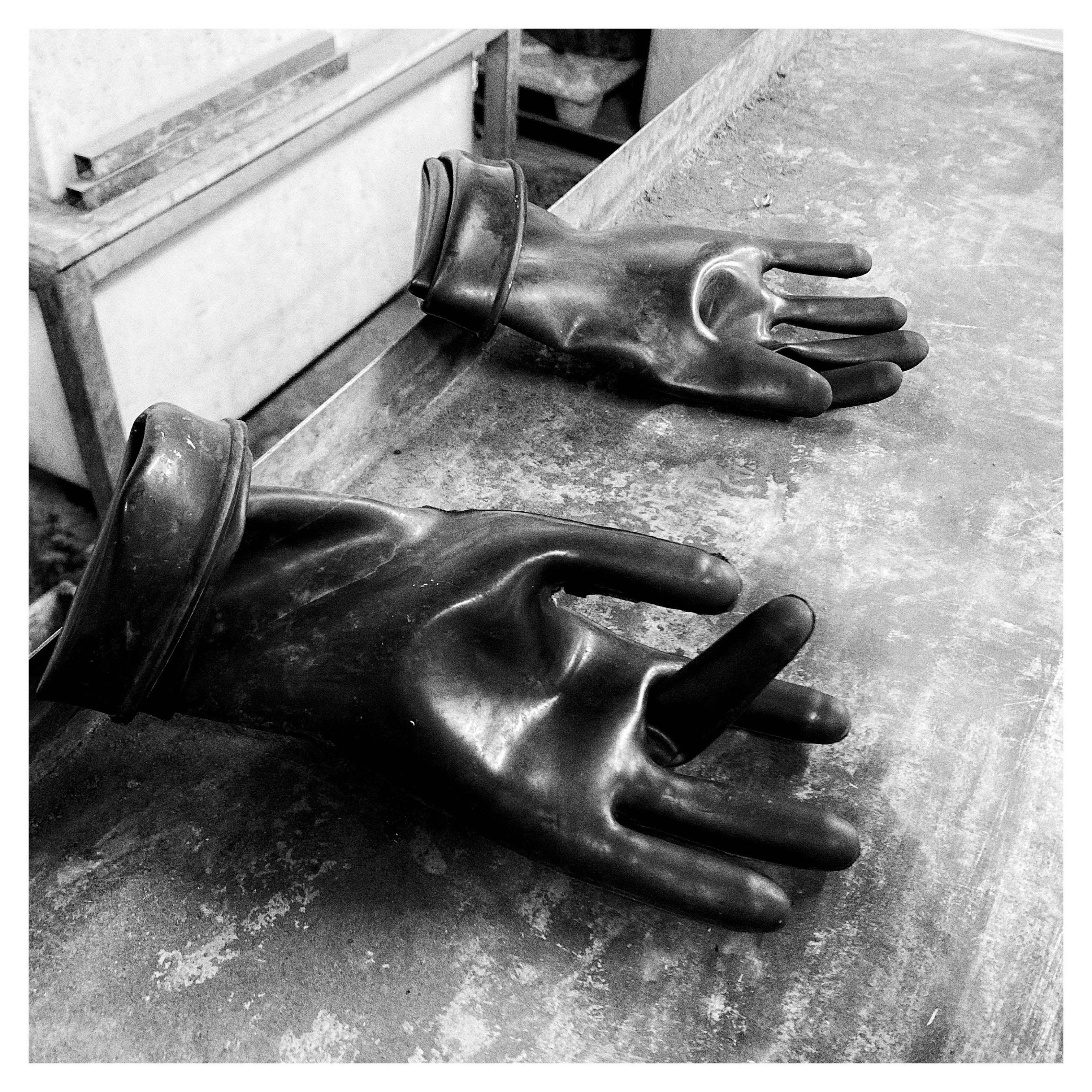 Two oversized rubber gloves lying on a worn worktable, one in the foreground with fingers splayed and one farther back, in a black-and-white workshop setting.
