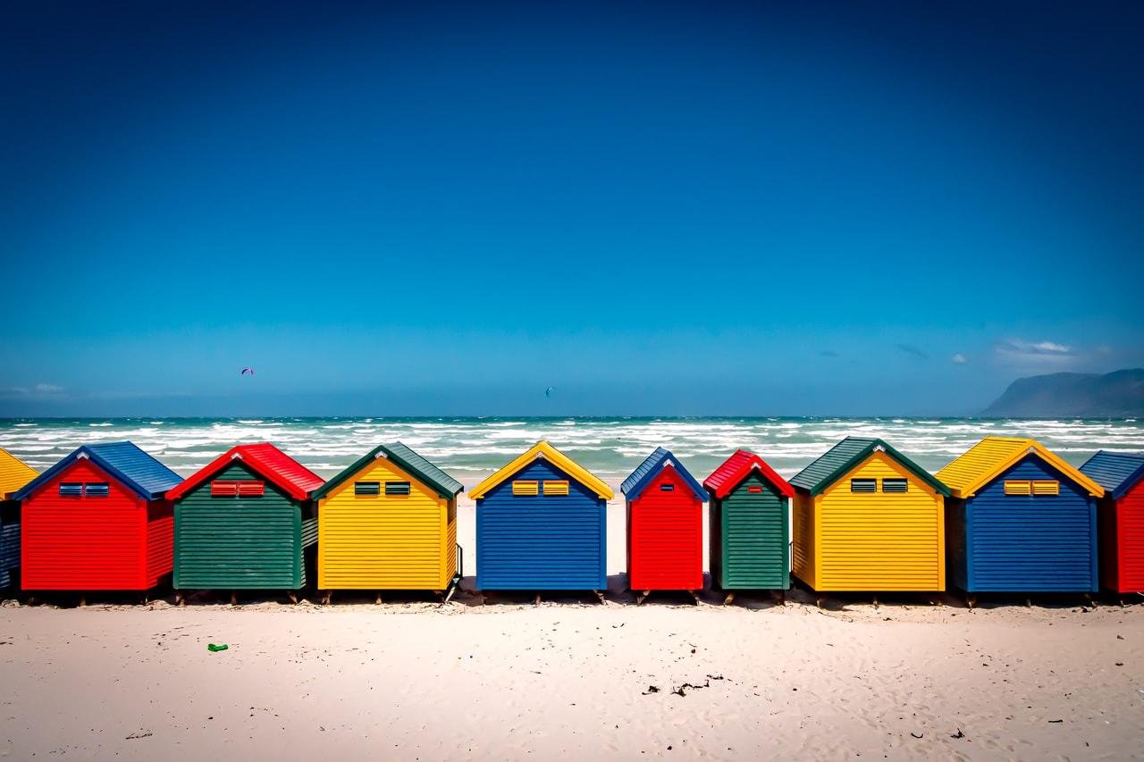 The image features a row of colorful beach huts on a sandy beach. The huts are painted in vibrant colors like blue, red, green, and yellow. In the background, there is a view of the ocean with waves and a clear blue sky.