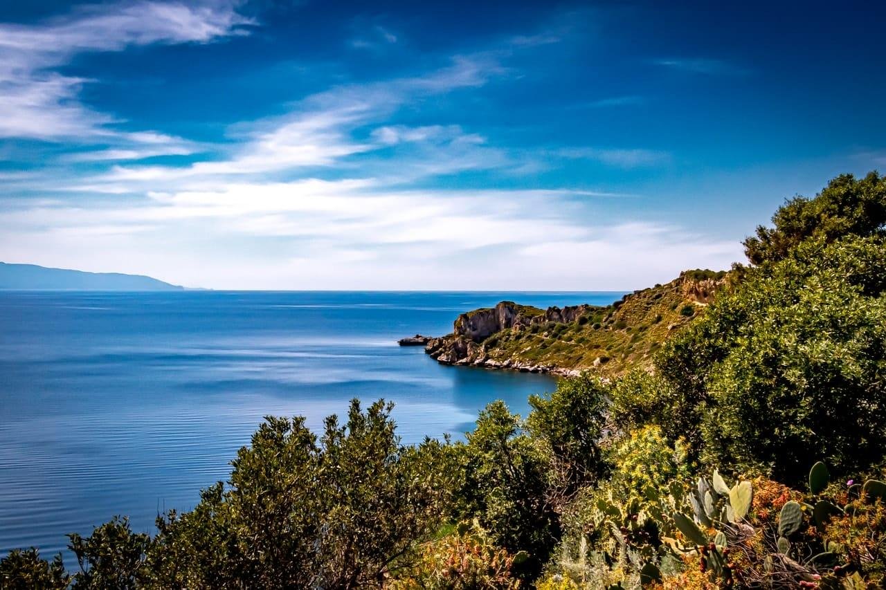 View from a hillside covered in shrubs and cactus overlooking a calm blue sea, with a rocky, green coastline curving to the right and distant land on the left under a bright sky with wispy clouds.
