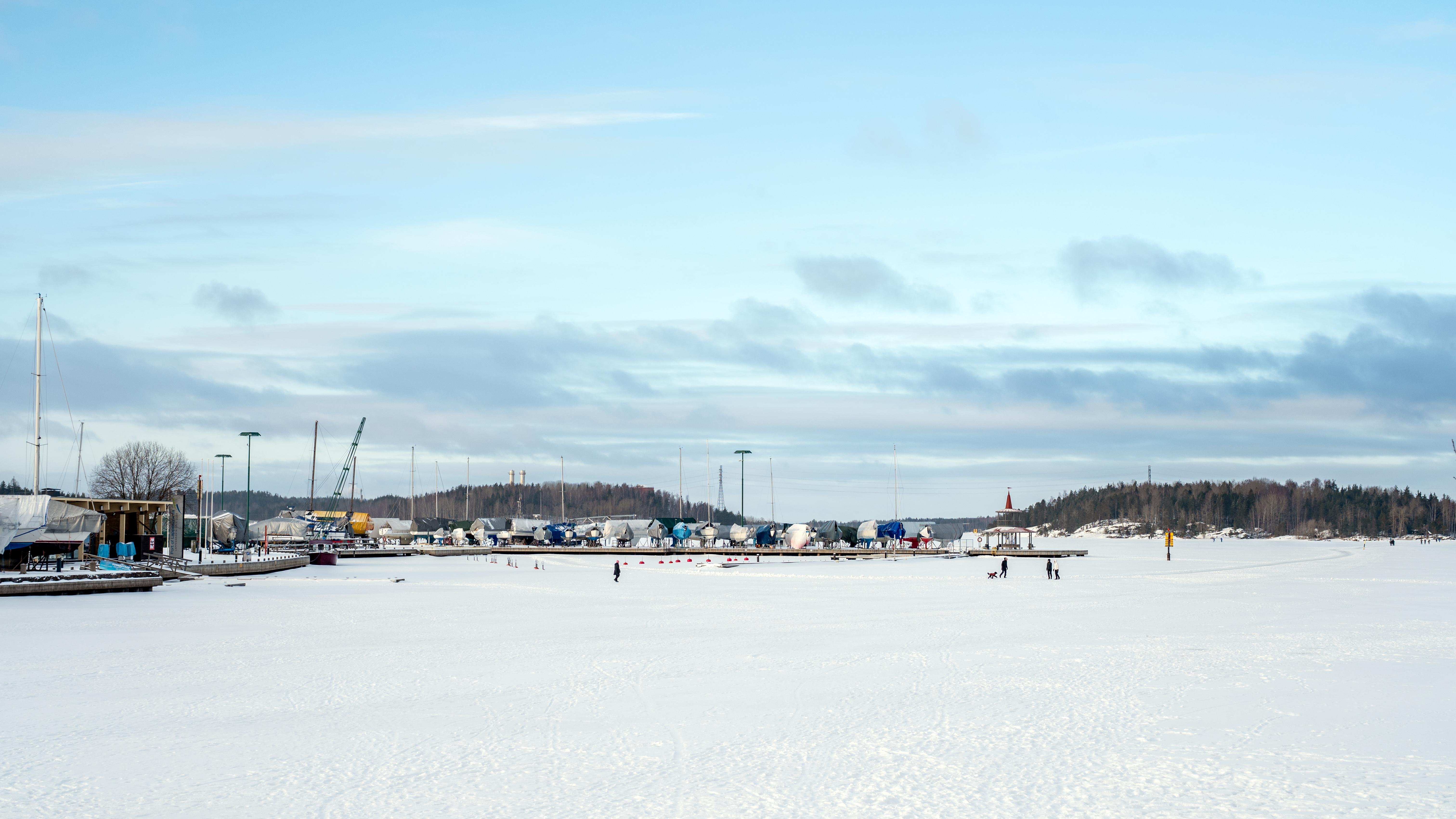 The image shows a wide, snowy landscape with a partially frozen body of water in the foreground. There are several people in the distance, possibly walking or ice skating. In the background, there are trees, buildings, and industrial equipment, like cranes, indicating a harbor or port area. The sky is mostly clear with some clouds.