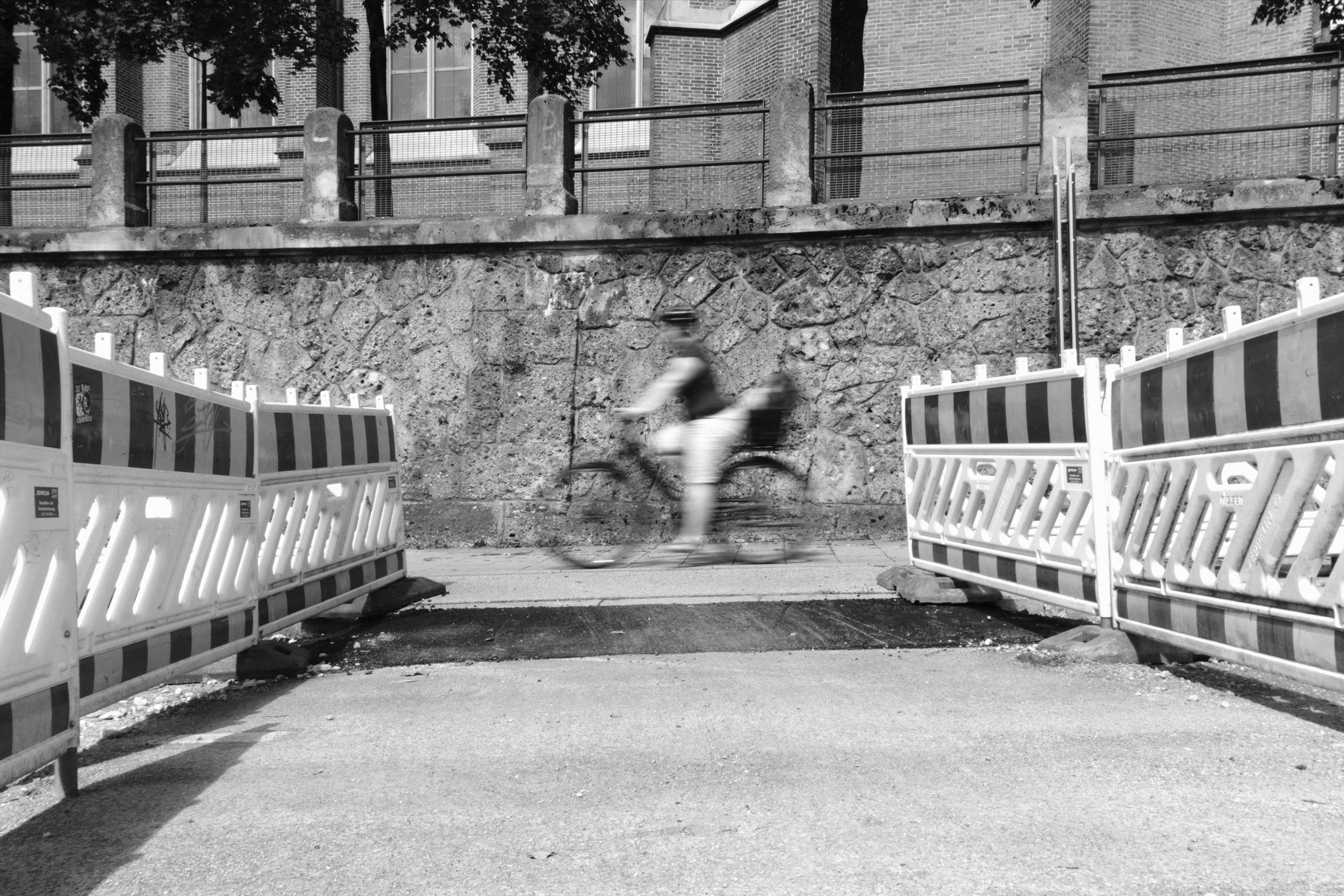 Black-and-white street scene with two rows of white construction barriers forming a corridor in the foreground; a motion-blurred cyclist rides across the center in front of a stone retaining wall with a metal fence and building windows above. The cyclist is posting the central opening of the corridor, with the construction barriers leading the view towards the cyclist.