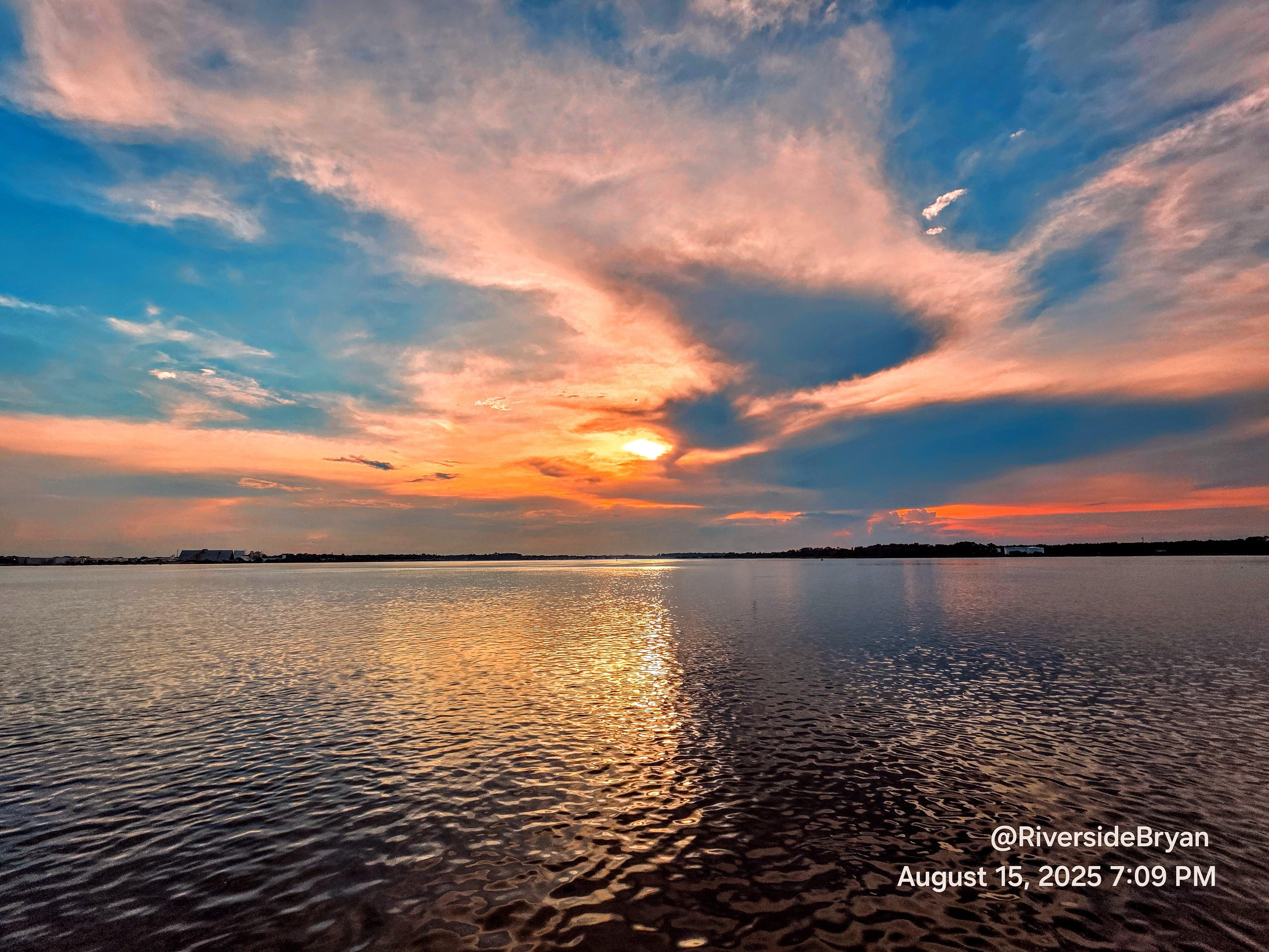 Sunset over a calm body of water with vibrant orange, pink, and blue clouds reflecting on the surface. The horizon line is low, with distant dark land visible. Text in the bottom right corner reads "@RiversideBryan August 15, 2025 7:09 PM".
