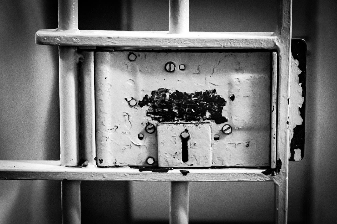 The image shows a close-up of an old prison cell door lock. The surface has peeling paint and visible screws, giving it a worn, aged appearance. The keyhole is prominently featured, reinforcing the look of a secure, sturdy lock. The image is in black and white, enhancing the texture and details.