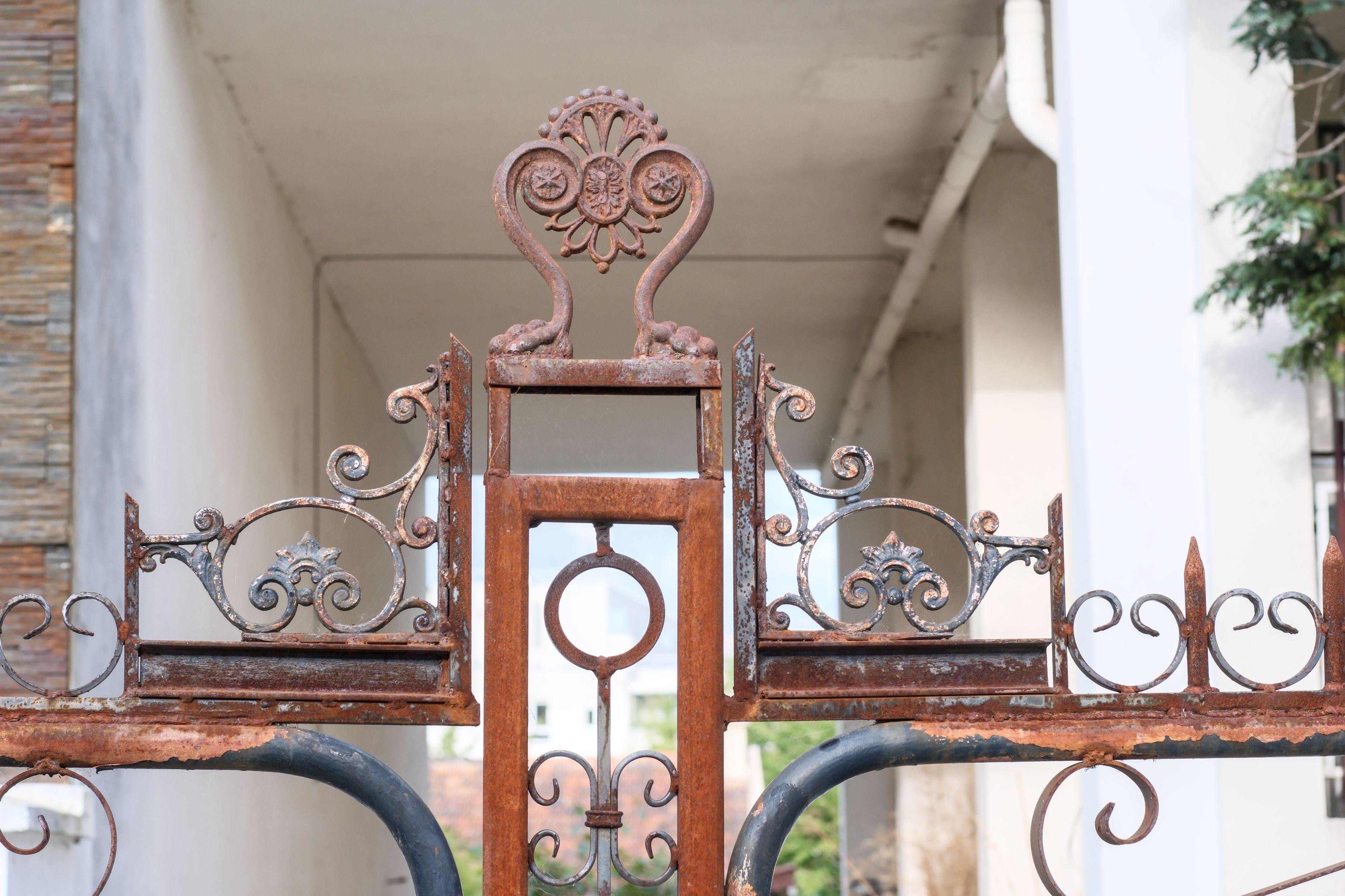 Photo format paysage, gros plan d’un ornement en haut d’un portail en métal rouillé. Du métal tout en courbes dans un style assez floral et végétal, difficile à décrire