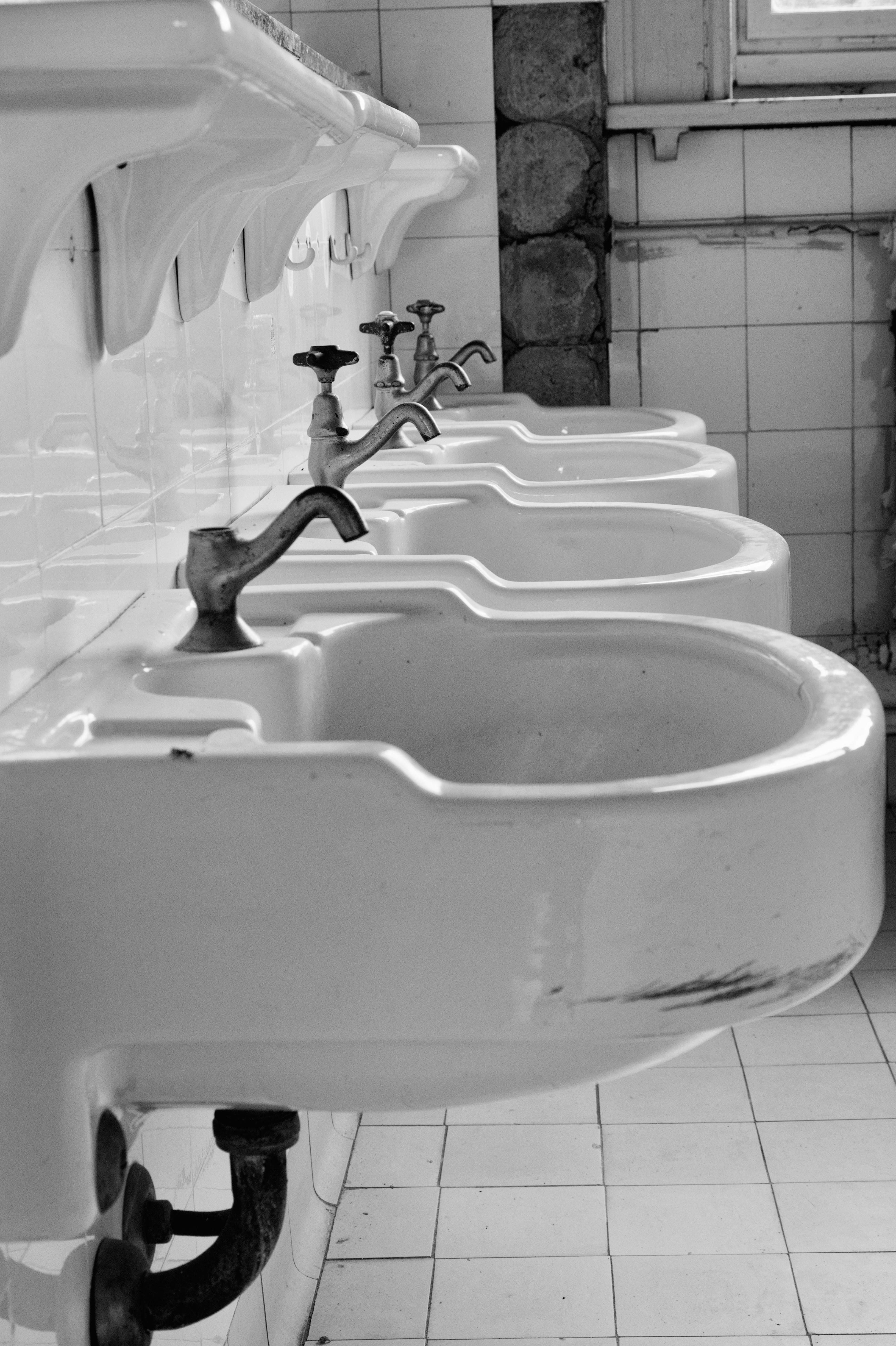 Row of vintage ceramic sinks with metal faucets mounted on a tiled wall. The floor is also tiled, and a window is visible in the background. Each sink is connected by visible plumbing pipes underneath. The scene is captured in black and white, giving a historical ambiance. 
### 
Reihe von Vintage-Keramikspülen mit Metallarmaturen, die an einer gefliesten Wand montiert sind. Der Boden ist ebenfalls gefliest, im Hintergrund ist ein Fenster zu sehen. Jedes Waschbecken ist durch sichtbare Rohrleitungen darunter verbunden. Die Szene ist in Schwarz-Weiß gehalten und vermittelt ein historisches Ambiente.