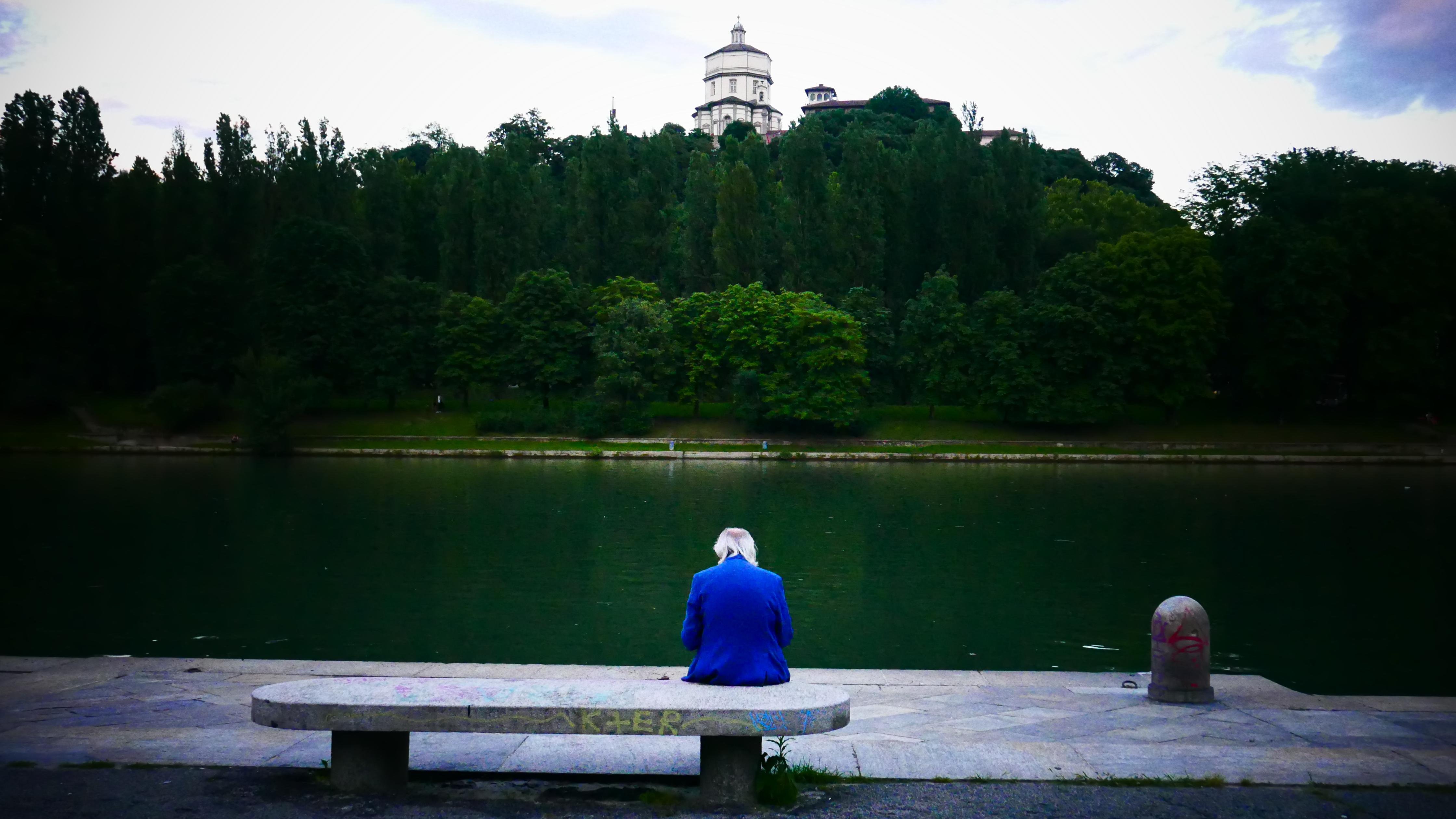 The image shows a person sitting on a stone bench by a body of water, facing away from the camera. The person is wearing a blue jacket. Across the water, there is a wooded area with a building with a dome on top of a hill or elevated area. The scene has a calm and serene atmosphere.