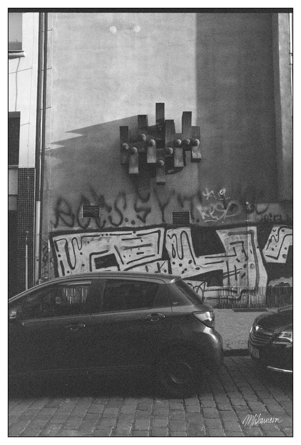Graffiti-covered wall featuring abstract design and text, located on a street with cobblestone pavement. Two parked cars are partially visible, one prominently in the foreground. A geometric metal structure is mounted above the graffiti on the building facade. Image is in black and white.