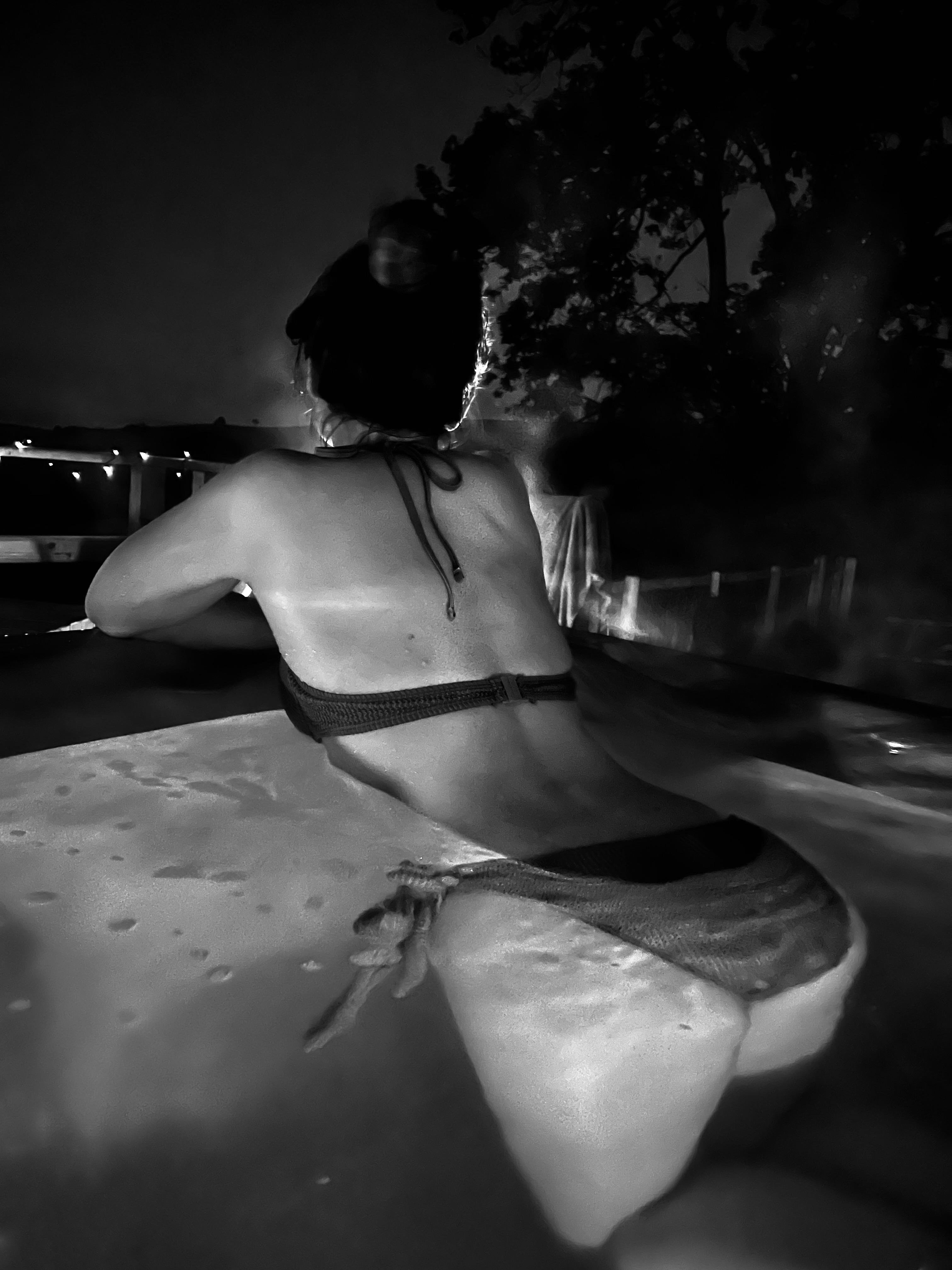 Rear view of a woman in a bikini leaning on the edge of an outdoor hot tub at night, with water droplets on the tub surface and a dimly lit fence and tree silhouette in the background. Photo is black and white.