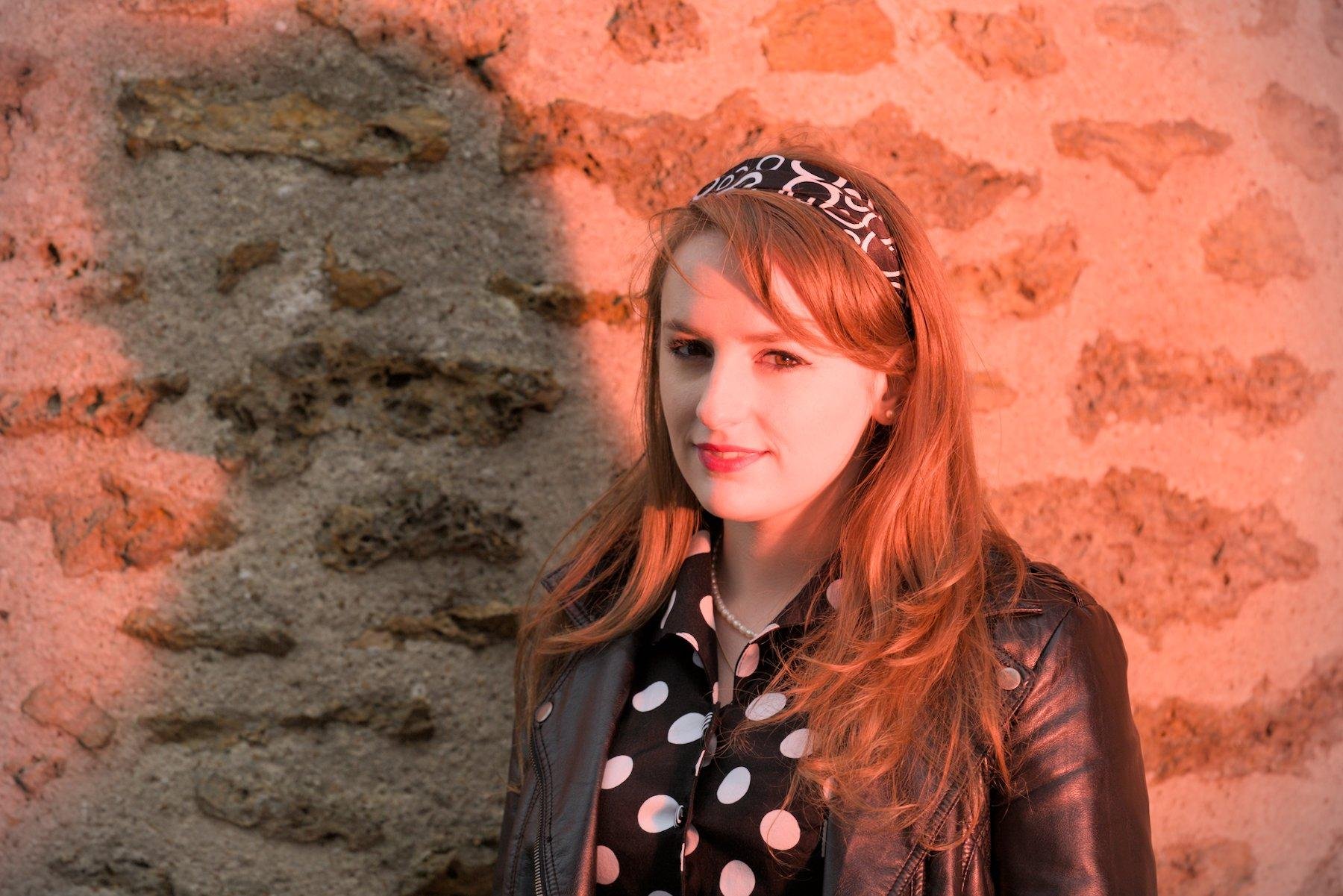 Portrait épaule d'une brune aux cheveux mi-longs portant une robe à point de pois noir et blanc, une veste en cuir noir et un serre-tête à motifs noir et blanc, debout à l'extérieur devant un mur en pierre, souriante et regardant vers la caméra. Son ombre est projetée sur le mur, dessinant son profil