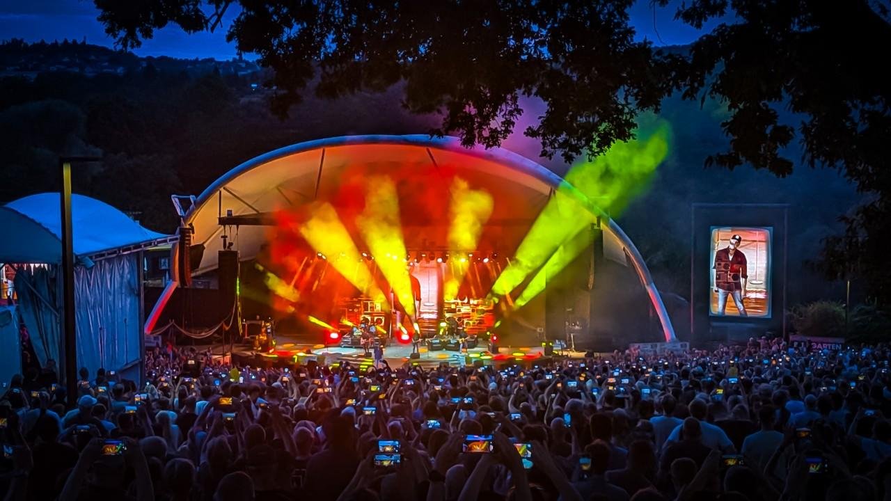 Outdoor concert at night with large crowd holding up phones. Stage illuminated by colorful lights with performers in front of a large, curved canopy. Bright yellow and orange beams highlight the area. Trees frame the scene, and a large screen on the right displays a performer.