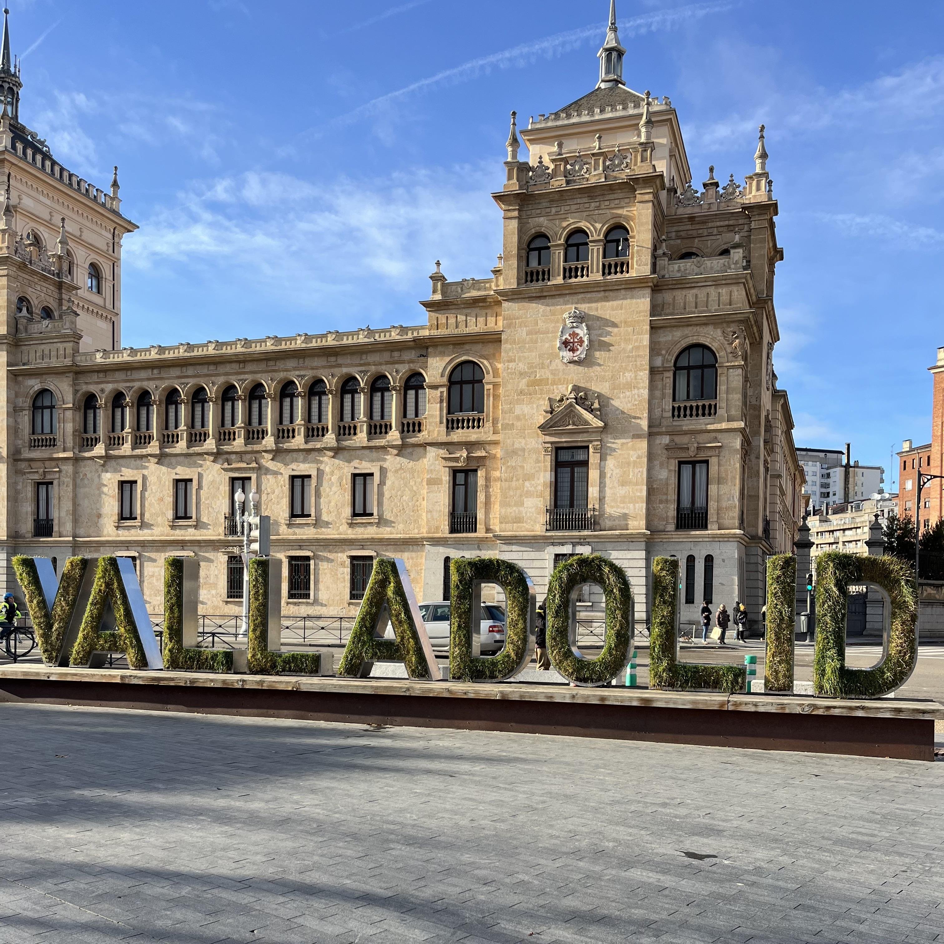 The image shows a building with architectural details and large letters in front spelling "VALLADOLID." The letters appear to be made from greenery or plants. The sky is clear and blue, indicating a sunny day.