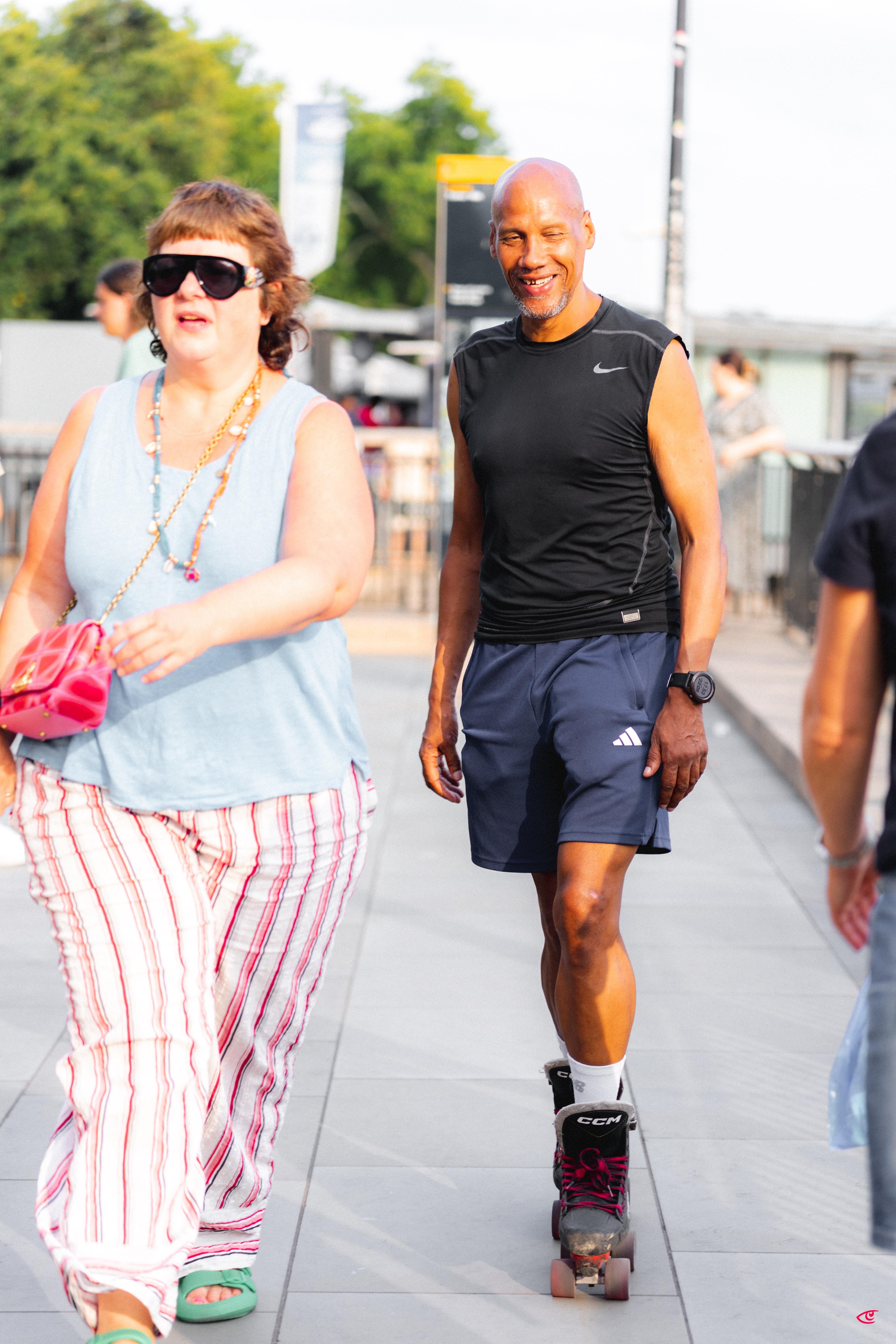 Two people on a city walkway: a woman in sunglasses wearing a light blue tank top and striped pants carrying a pink purse in the foreground, and a man in a black sleeveless shirt and navy shorts rollerblading behind her; trees, railings, and other pedestrians in the background.